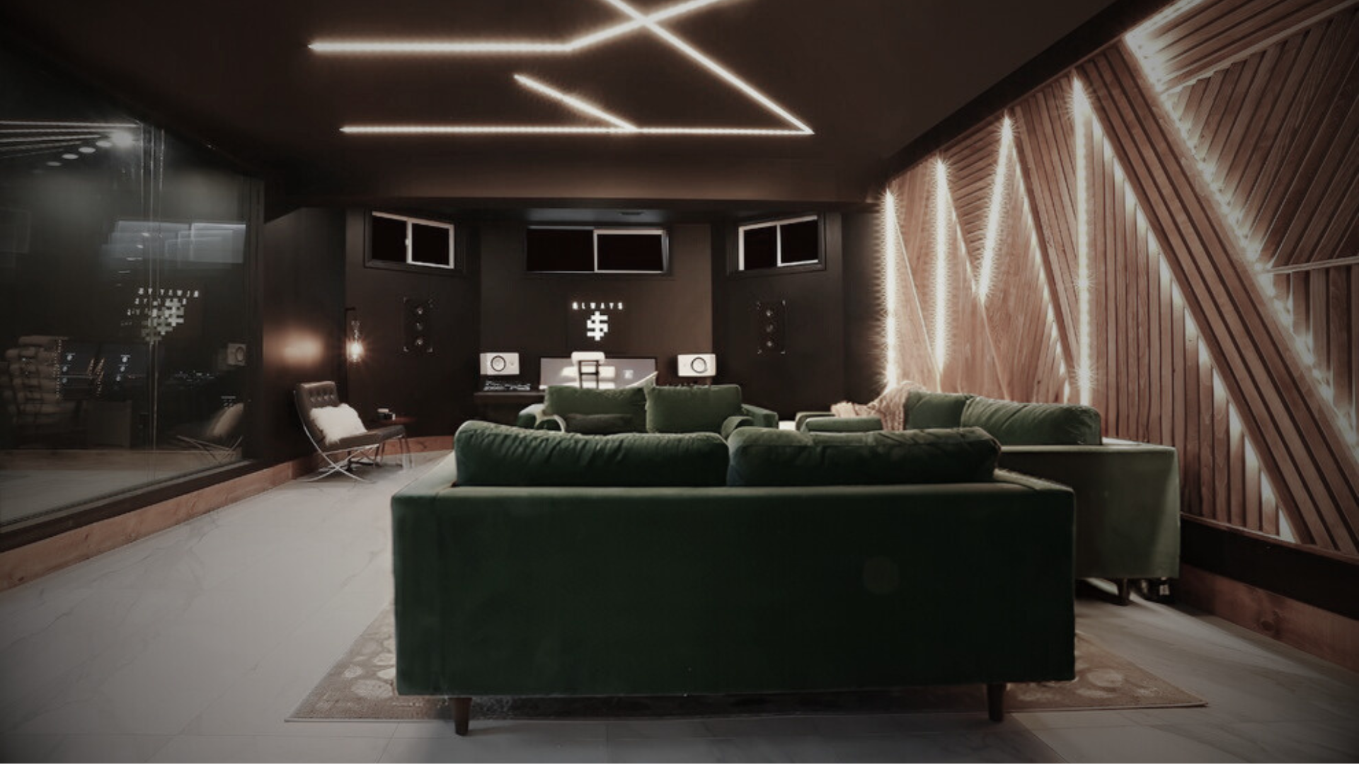 A modern recording studio with dark walls, green velvet sofas, and wooden accents illuminated by LED lighting.