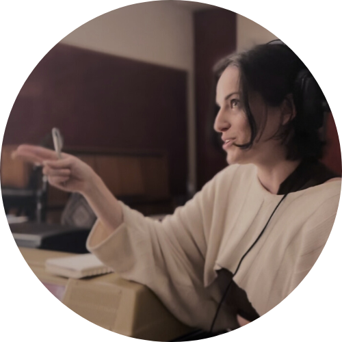 A woman with dark hair, gesturing with her right hand, sitting at a desk with papers, in an indoor setting.
