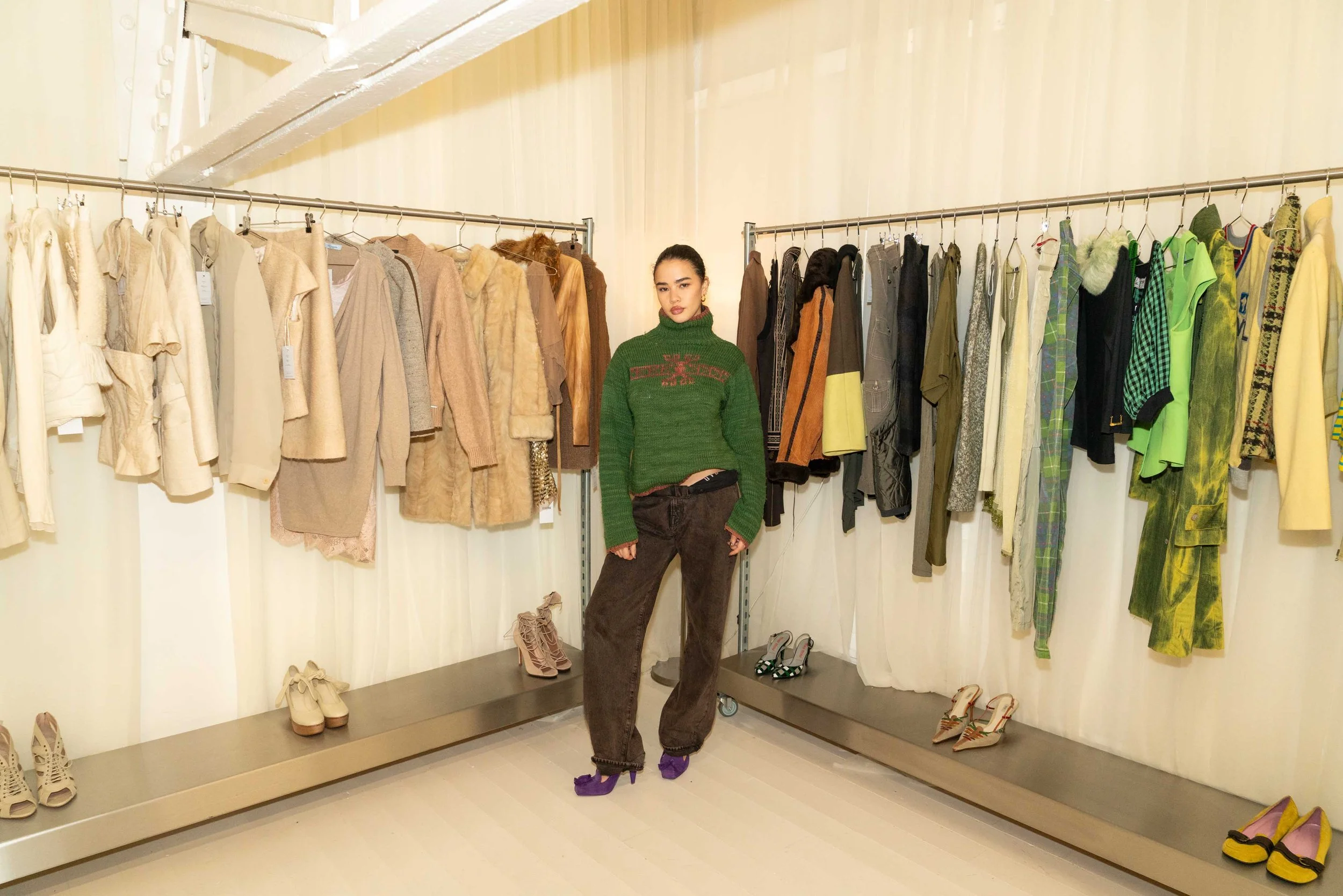 A woman standing in a clothing store with racks of various colorful jackets and shoes behind her.