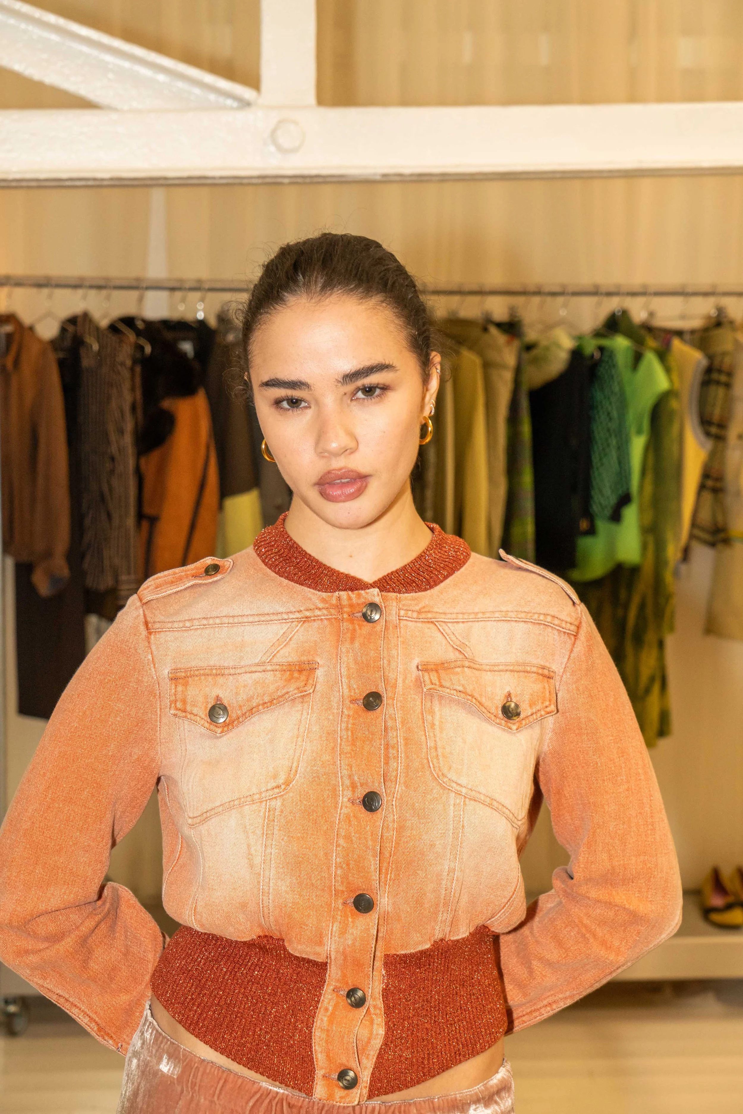 A woman wearing an orange denim jacket with a red waistband, standing in front of a clothing rack with various colored garments.