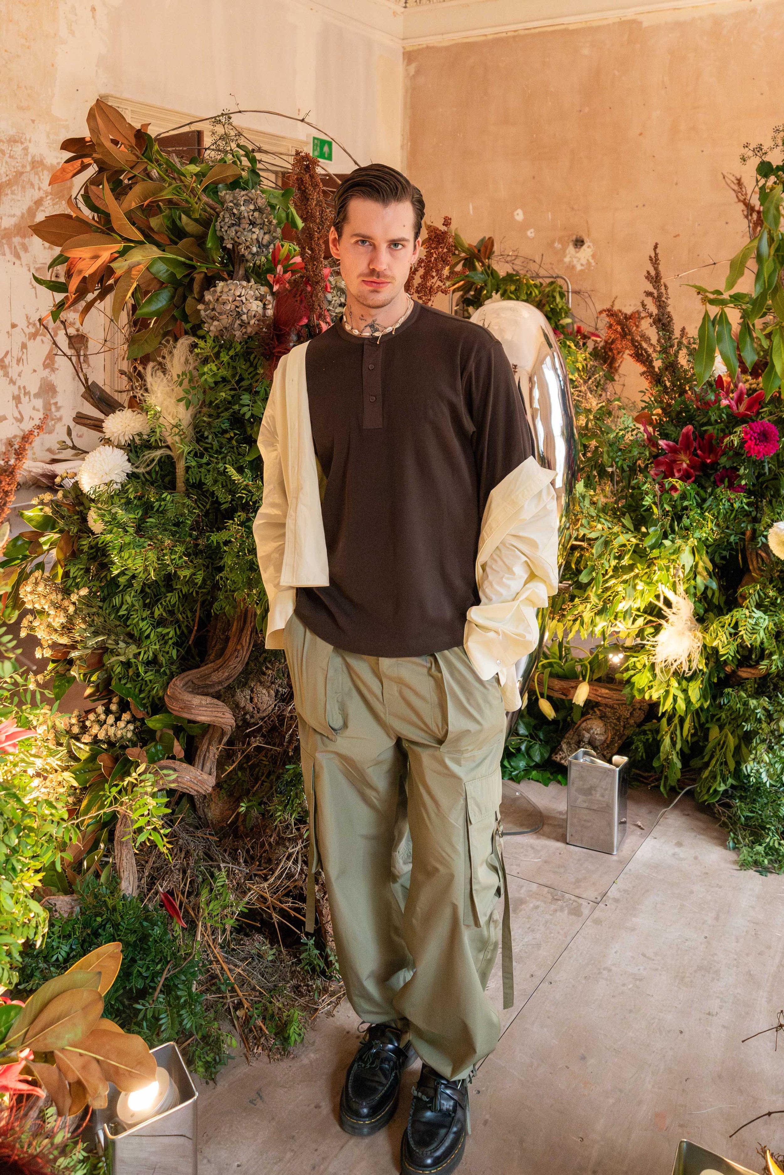A young man standing in front of a floral display with green plants, flowers, and decorative elements in an indoor setting.