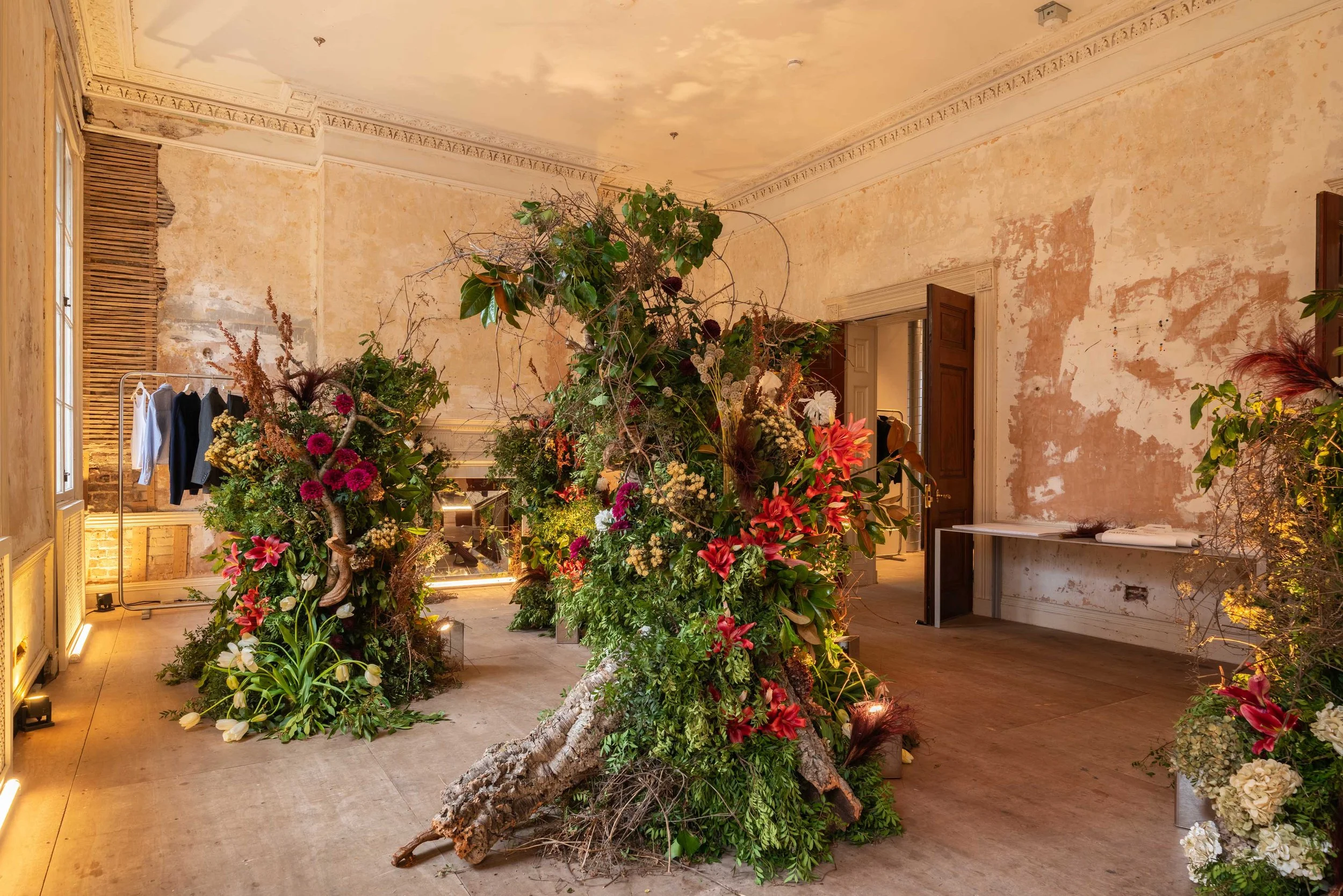 An indoor room with weathered walls, a large floral installation with greenery and assorted flowers in the center, and a clothing rack with hanging clothes on the left side.