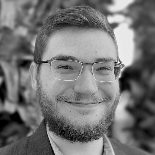 Black and white portrait of a young man with a beard, smiling at the camera, seated indoors with plants and office decor in the background.