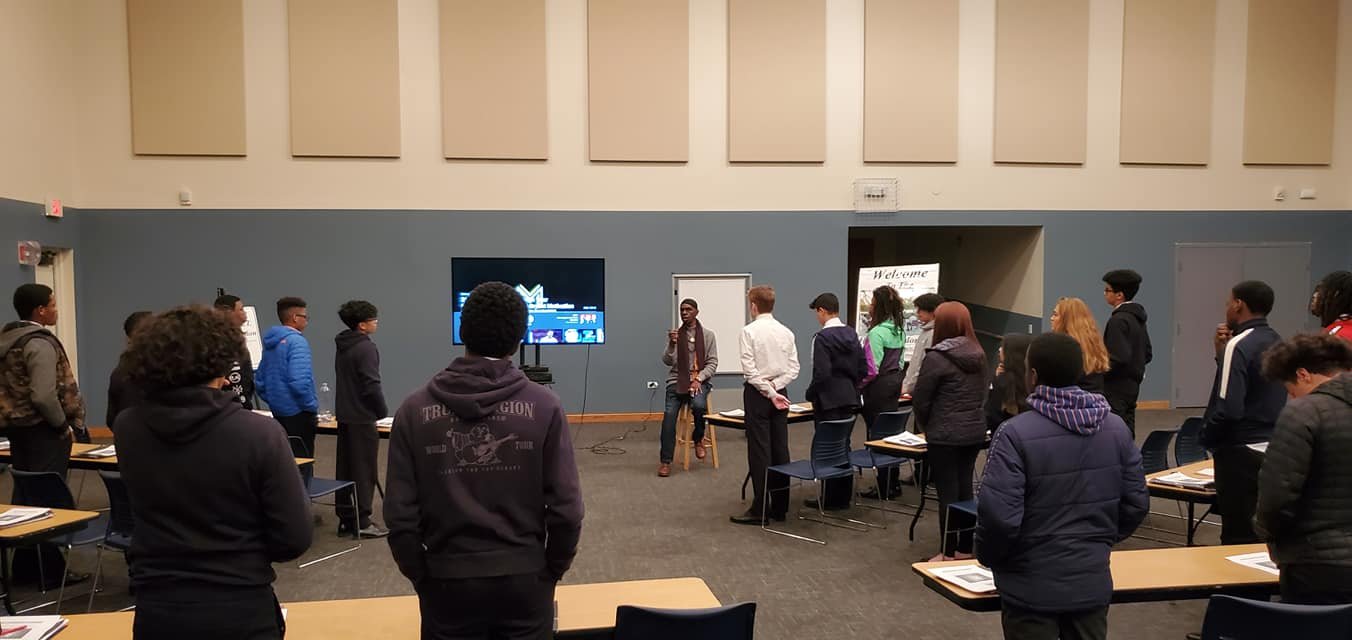 A group of people standing in a meeting room for Mentoring session with Bhagwan Ra Afrika. The speaker is at the front near a screen and appears to be addressing the group. Chairs and tables are arranged around the room.