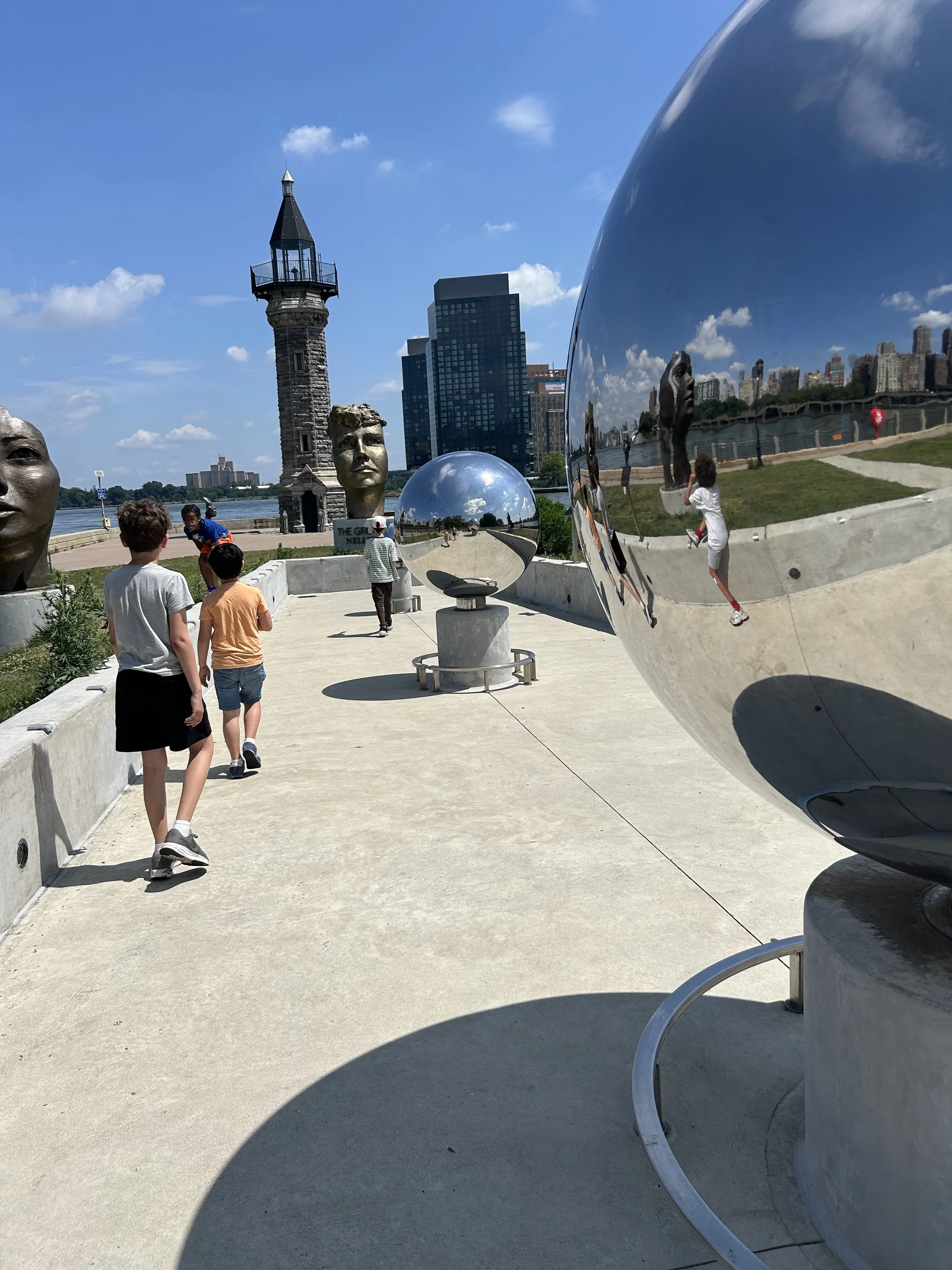 People walking at the Chicago Picasso sculpture installation near the river, with large metallic spheres and busts in the foreground, a lighthouse, and the city skyline in the background on a sunny day.