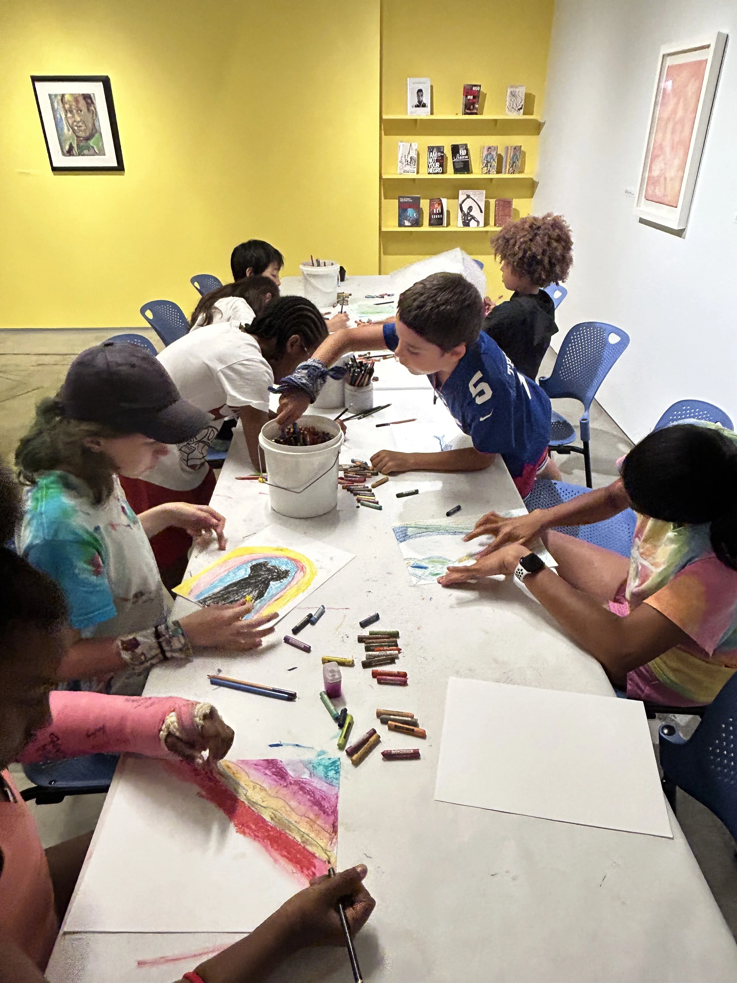 Children participating in an art activity at a long table, drawing and coloring with various art supplies, inside a colorful room with yellow and white walls, bookshelves, and framed artwork.