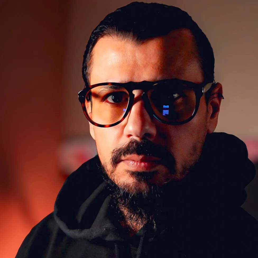 A man with dark hair, wearing glasses, and a beard, looking directly at the camera.