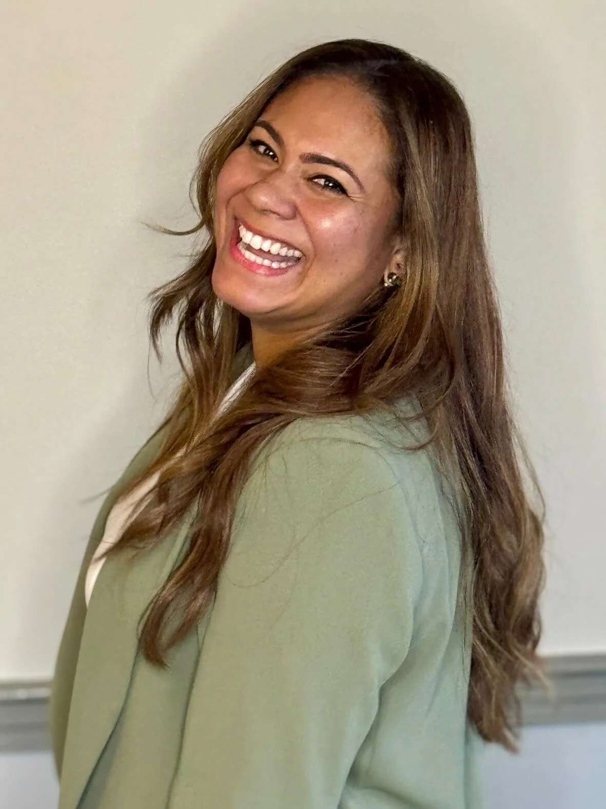 Ellen Bellamy, co-house manager, with long, wavy, brown hair and earrings, smiling and wearing a green blazer, standing against a plain light-colored background.