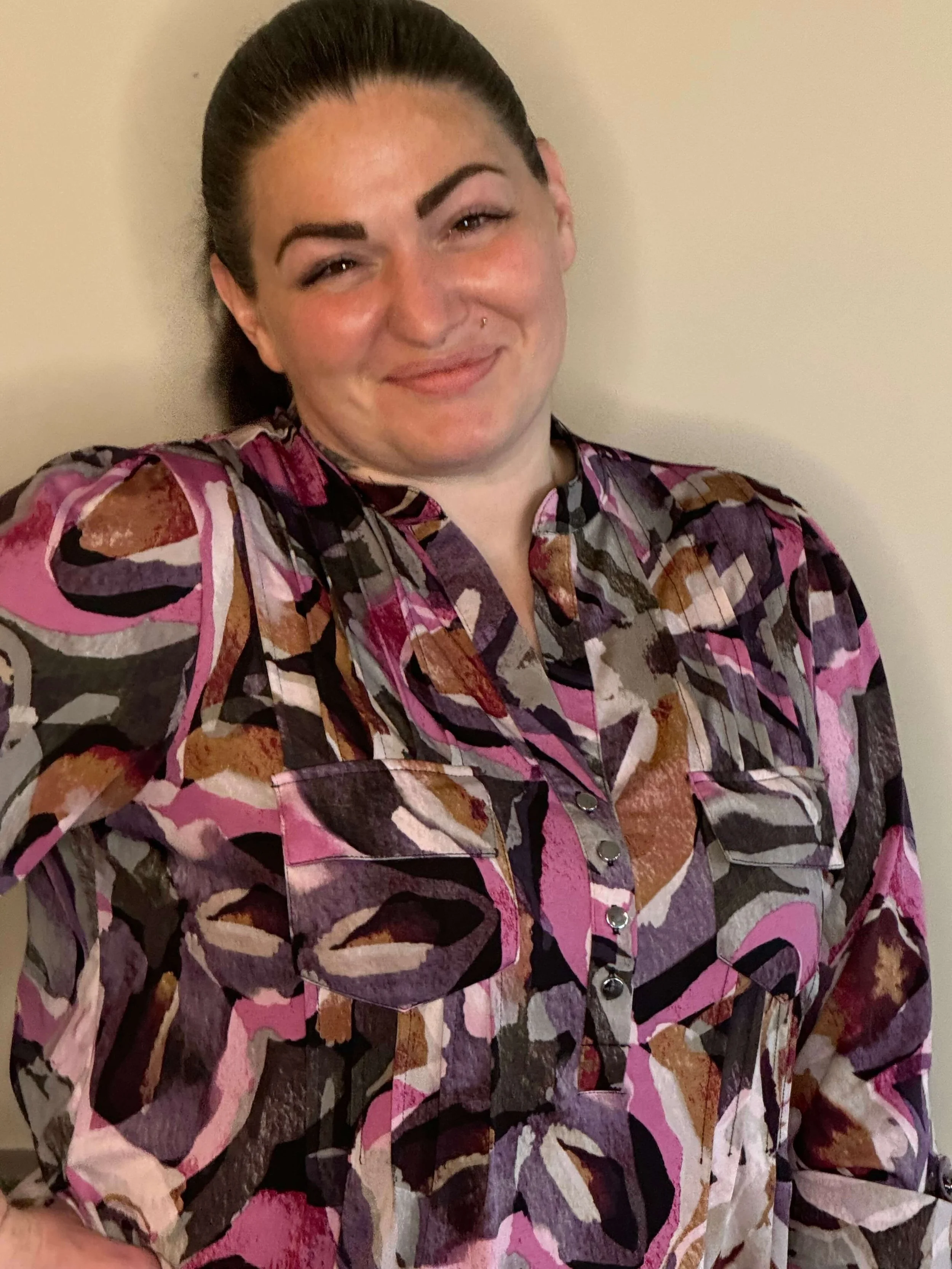 Moriah Beatrice, survivor advocate, with dark hair pulled back wearing a colorful, patterned shirt, smiling and standing against a plain wall.
