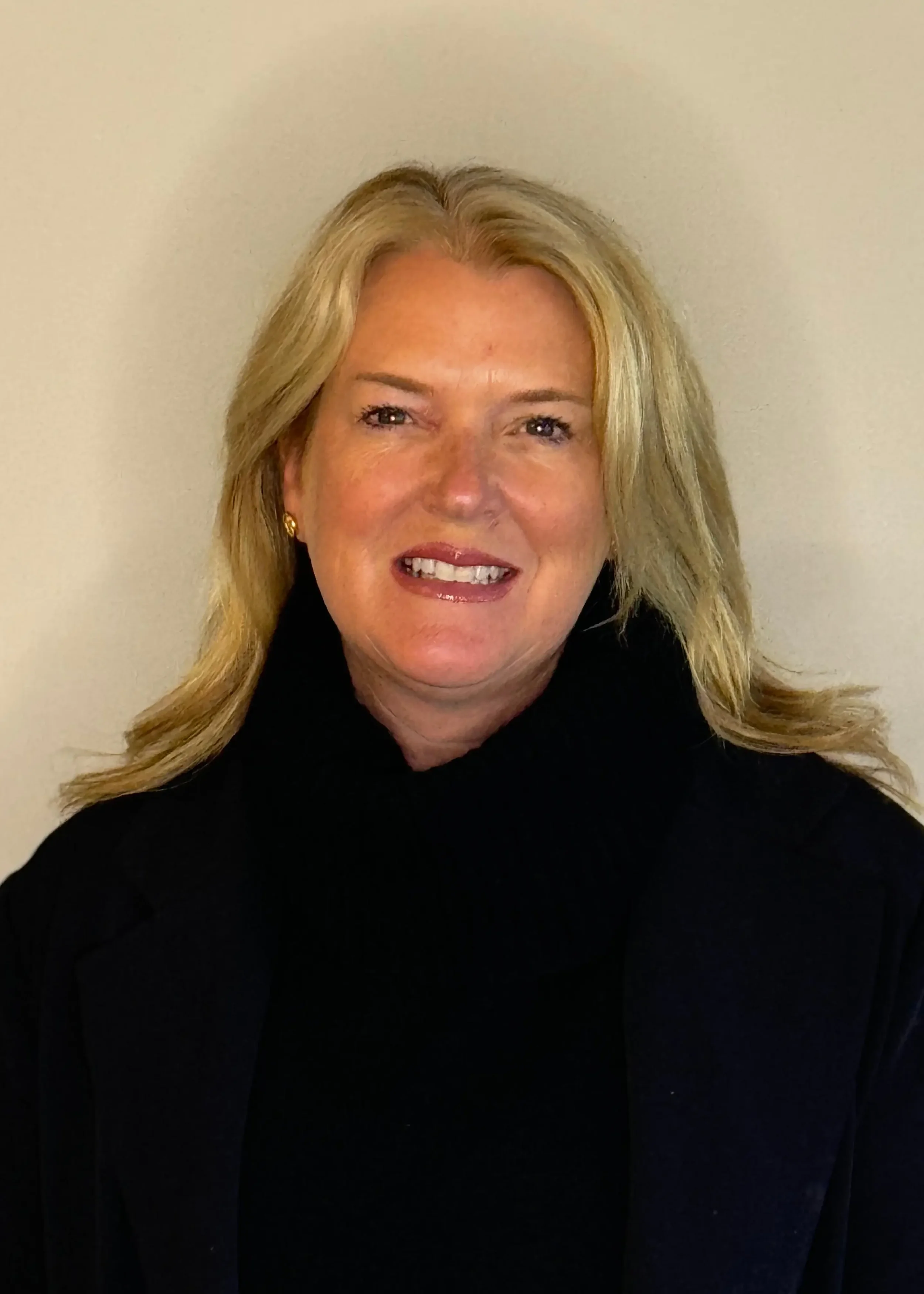 Close-up of Ali Boak, executive director emeritus, with blonde hair smiling, wearing a black top and earrings, standing against a plain white wall.