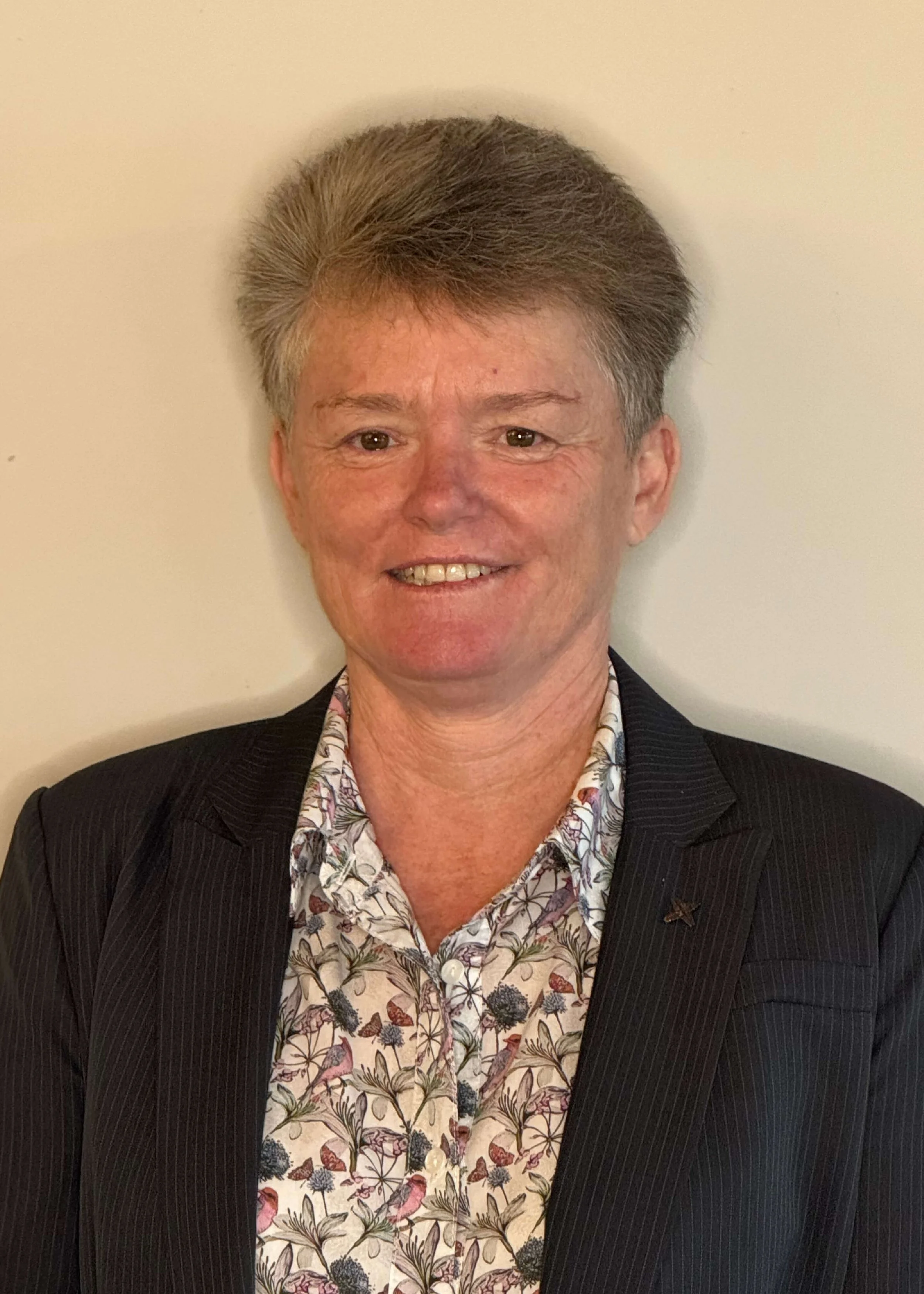 Sister Kate O'Neill, host community member, with short, gray hair styled upward, smiling, wearing a dark pinstripe blazer over a floral-patterned blouse, standing against a plain light-colored wall.