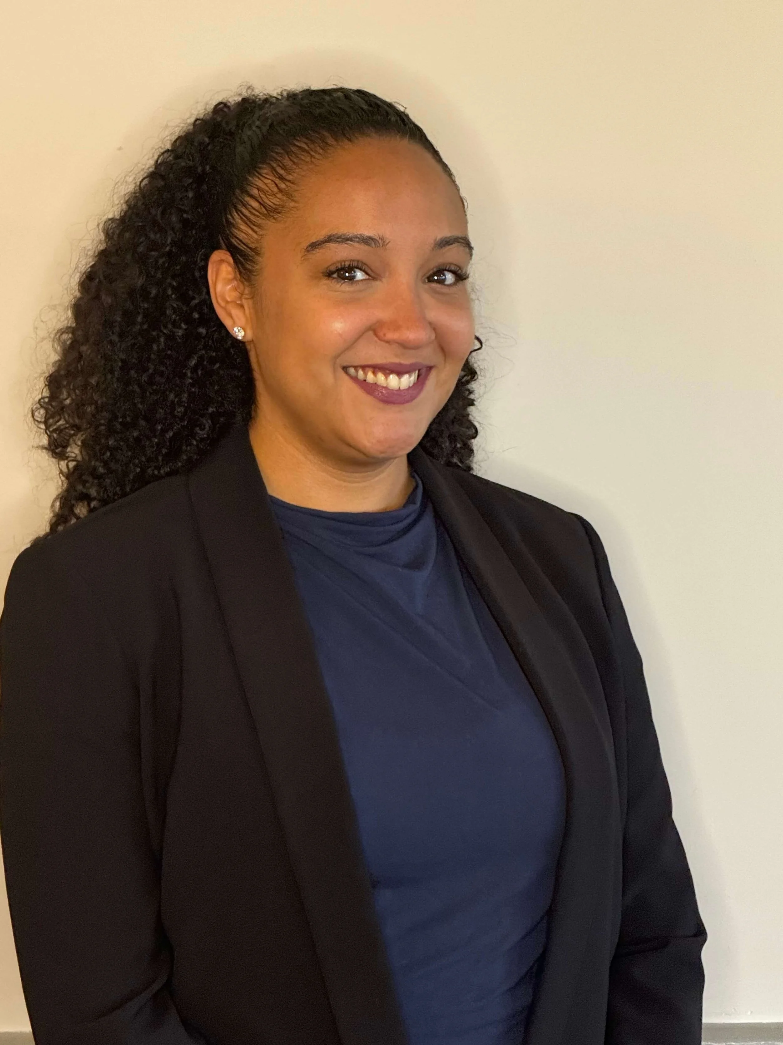 Josephine Crisostomo, director of people and operations, with curly black hair styled in a high ponytail, wearing a dark blazer and a blue top, smiling in front of a plain white wall.