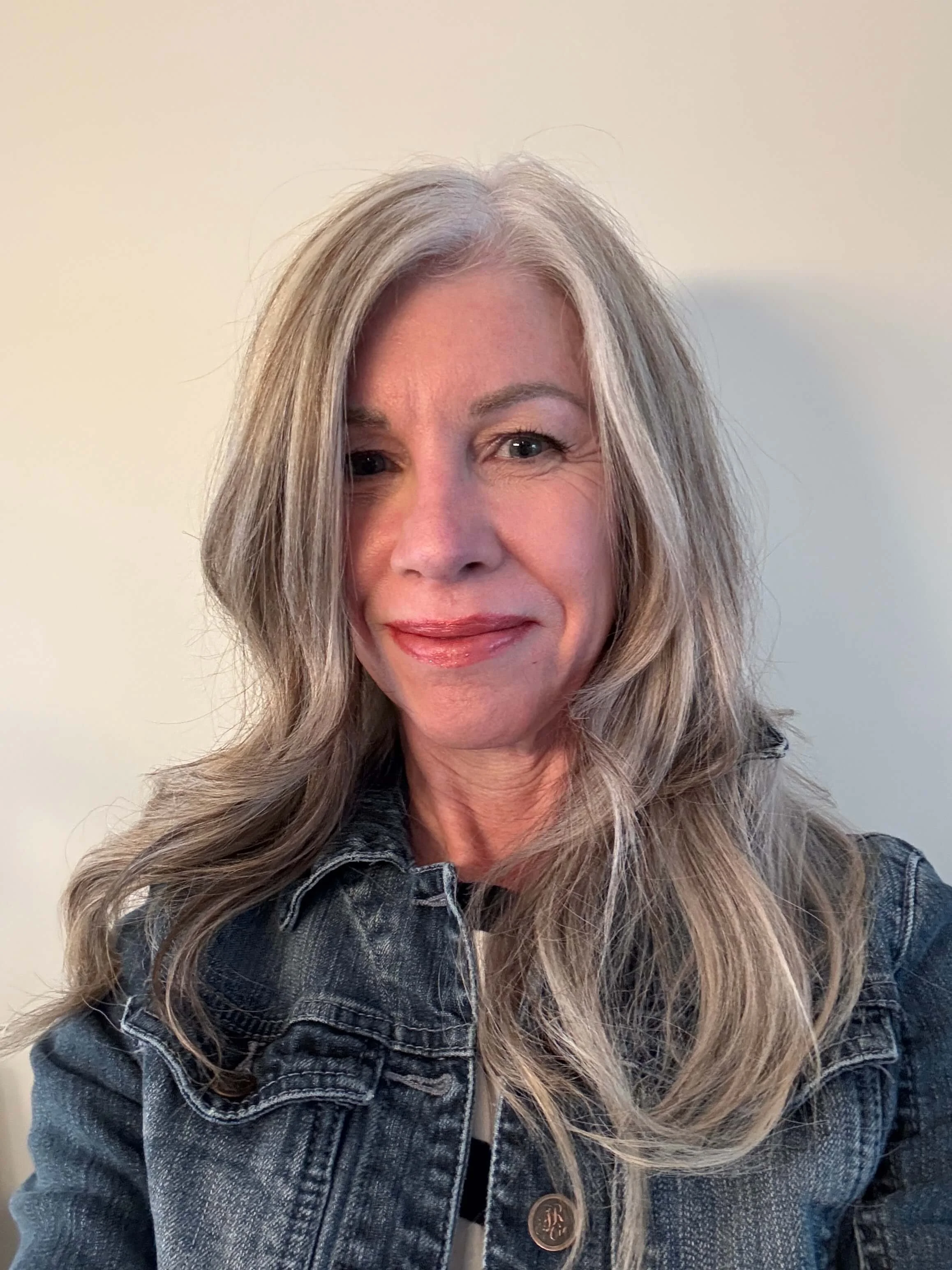 Julianne Will, marketing and communications specialist, with long, wavy white hair and light skin, smiling and wearing a denim jacket, standing against a plain light-colored background.