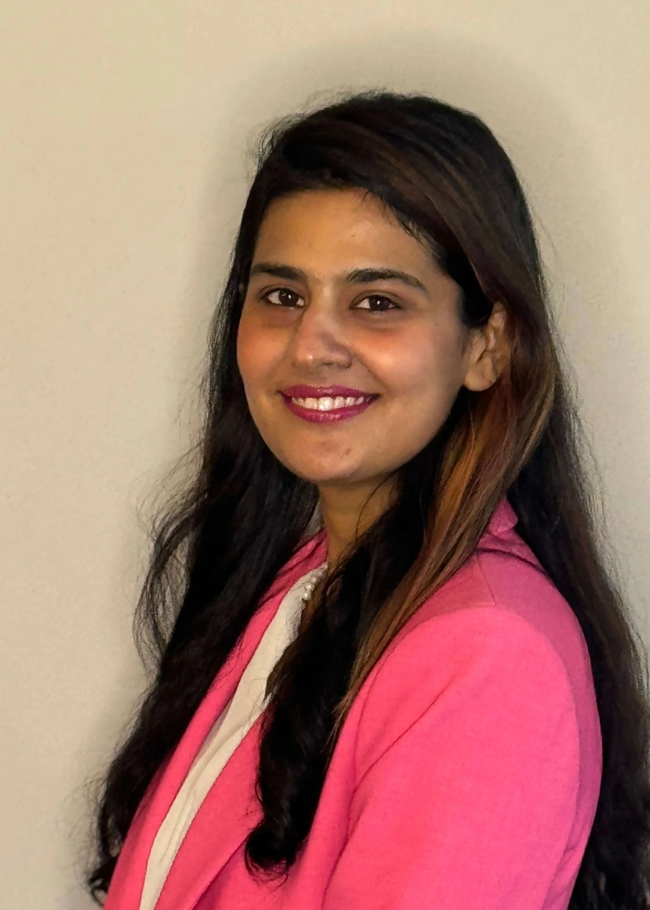Laraib Anjum, co-house manager, with dark, styled hair and a warm smile, wearing a pink garment with a cream-colored scarf, standing against a plain, light-colored wall.