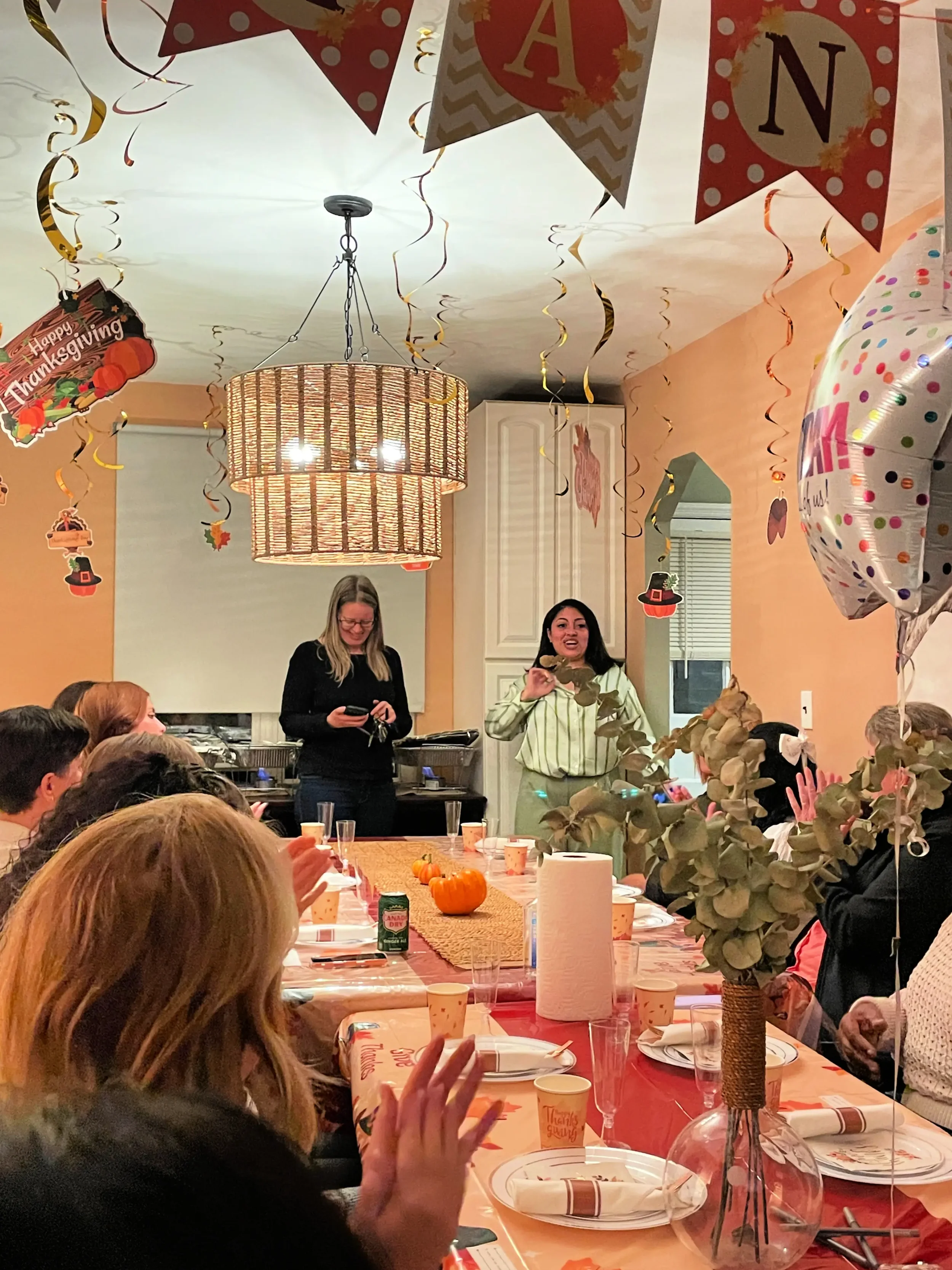 People gathered around a table for a Thanksgiving dinner celebration. Decorations include balloons, streamers, pumpkins, and a banner with the word 'GOBBLE'. Two women are standing and speaking to the group, one holding a microphone. The table is set with plates, napkins, cups, and a centerpiece with a floral arrangement.