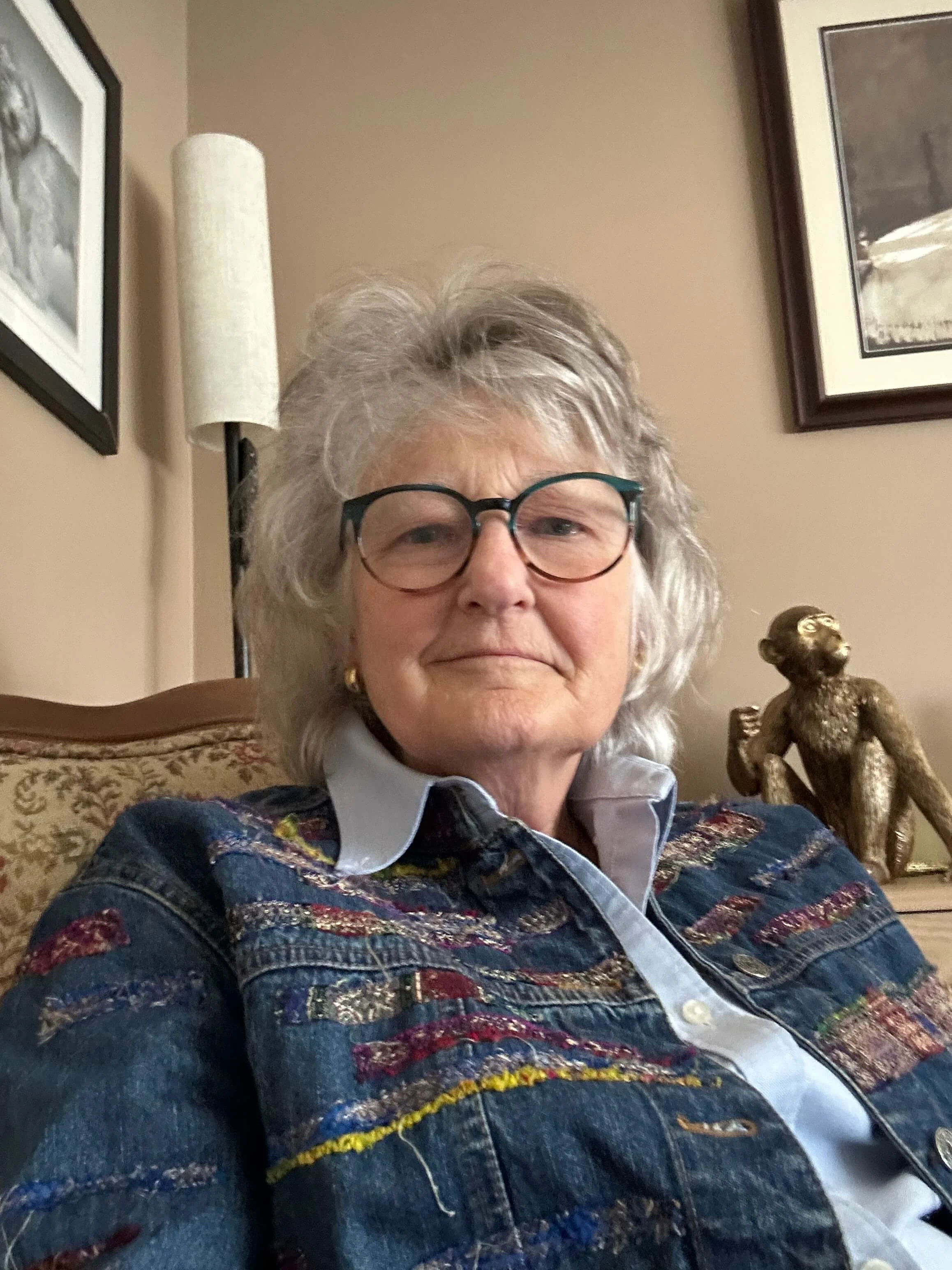 Mary McMahon, executive director, with gray hair, wearing glasses and a colorful embroidered jean jacket, is sitting on a patterned couch in a room with beige walls, framed artwork, and a decorative sculpture of a sitting woman in the background.