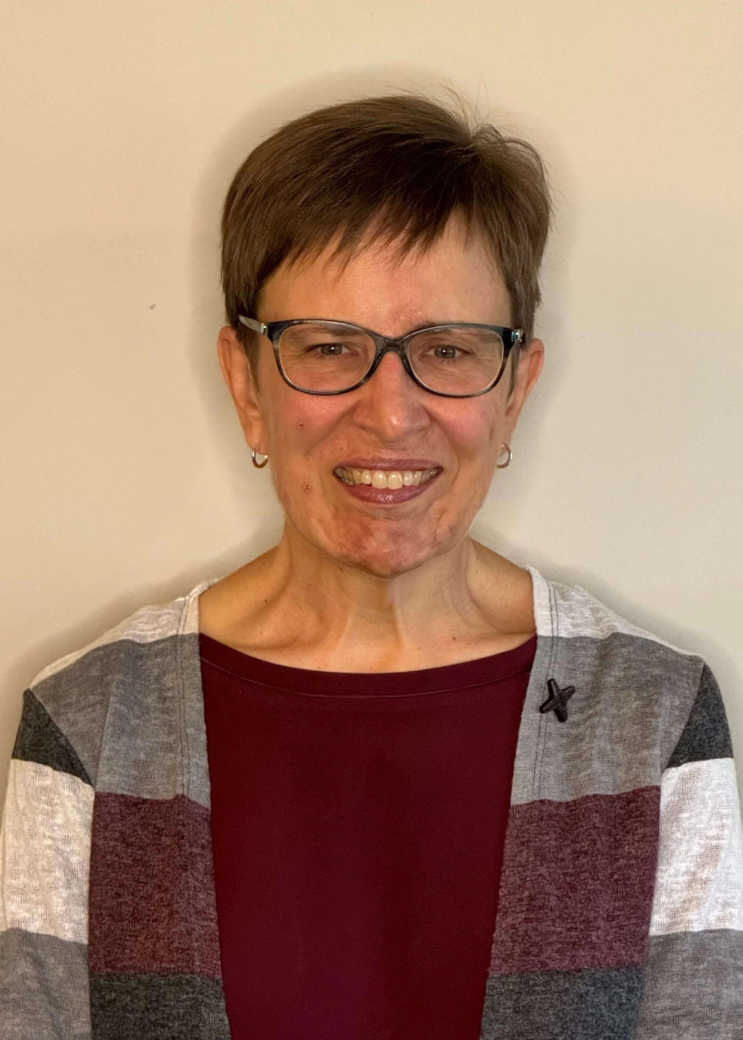 Sister Claudia Stecker, host community member, with short brown hair and glasses smiling, wearing a color-blocked sweater with maroon, light gray, and dark gray sections, against a plain white wall background.