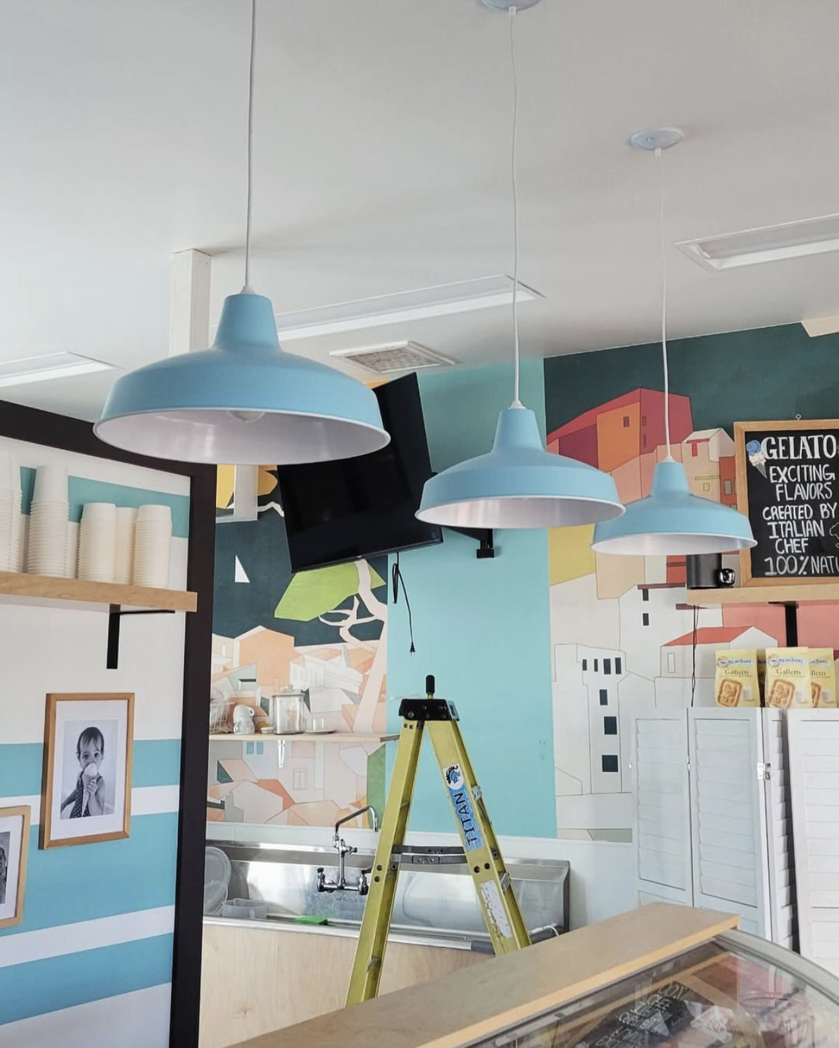 Interior of a gelato shop with blue hanging lights in a custom lighting job done by Marmolejo Electric, a colorful mural, a TV, a yellow ladder, framed pictures, and a menu on the wall in Solvang. 