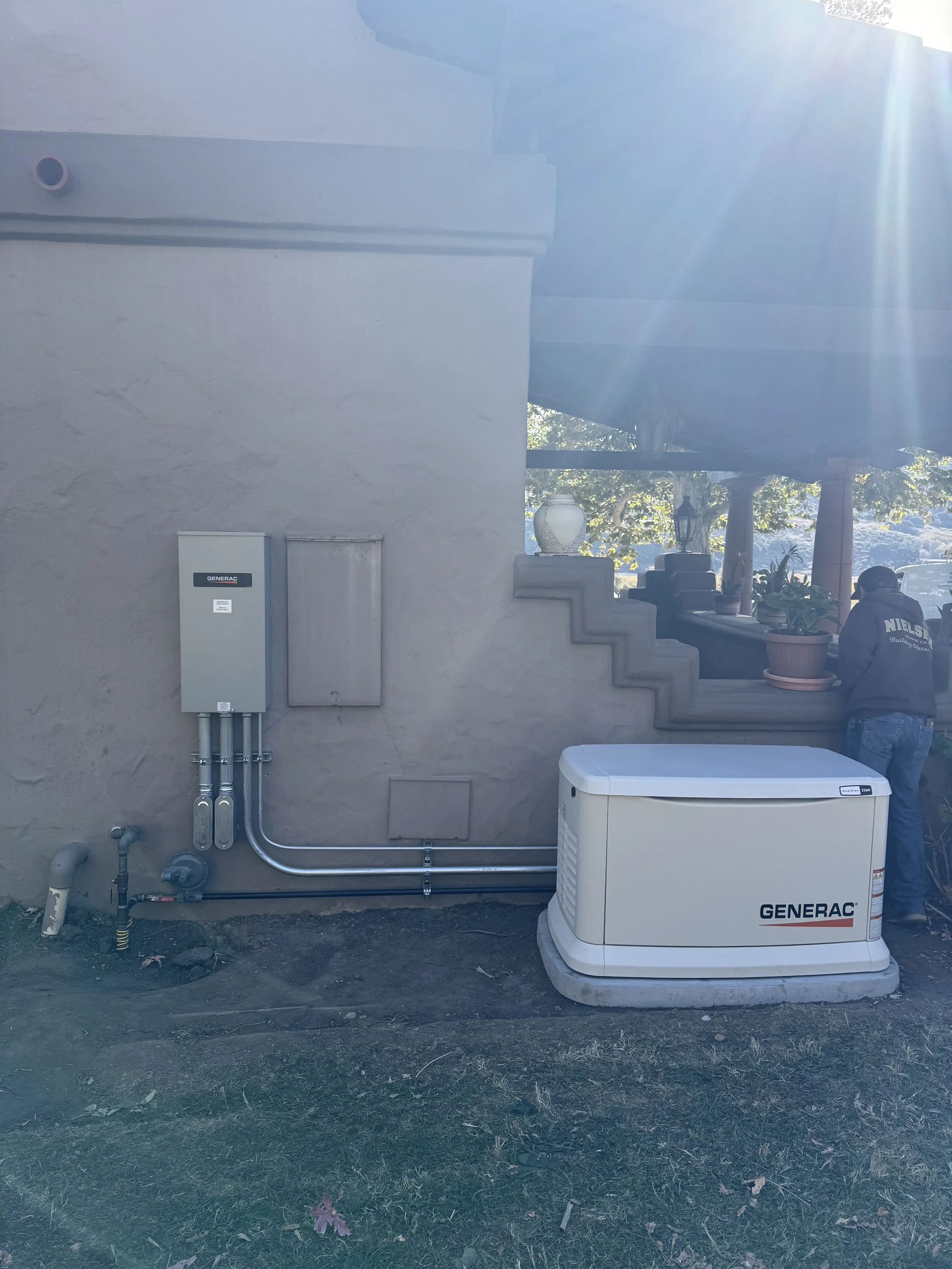 Home generator (Generac) installed next to a building wall with electrical conduits and utility connections, outdoor setting.