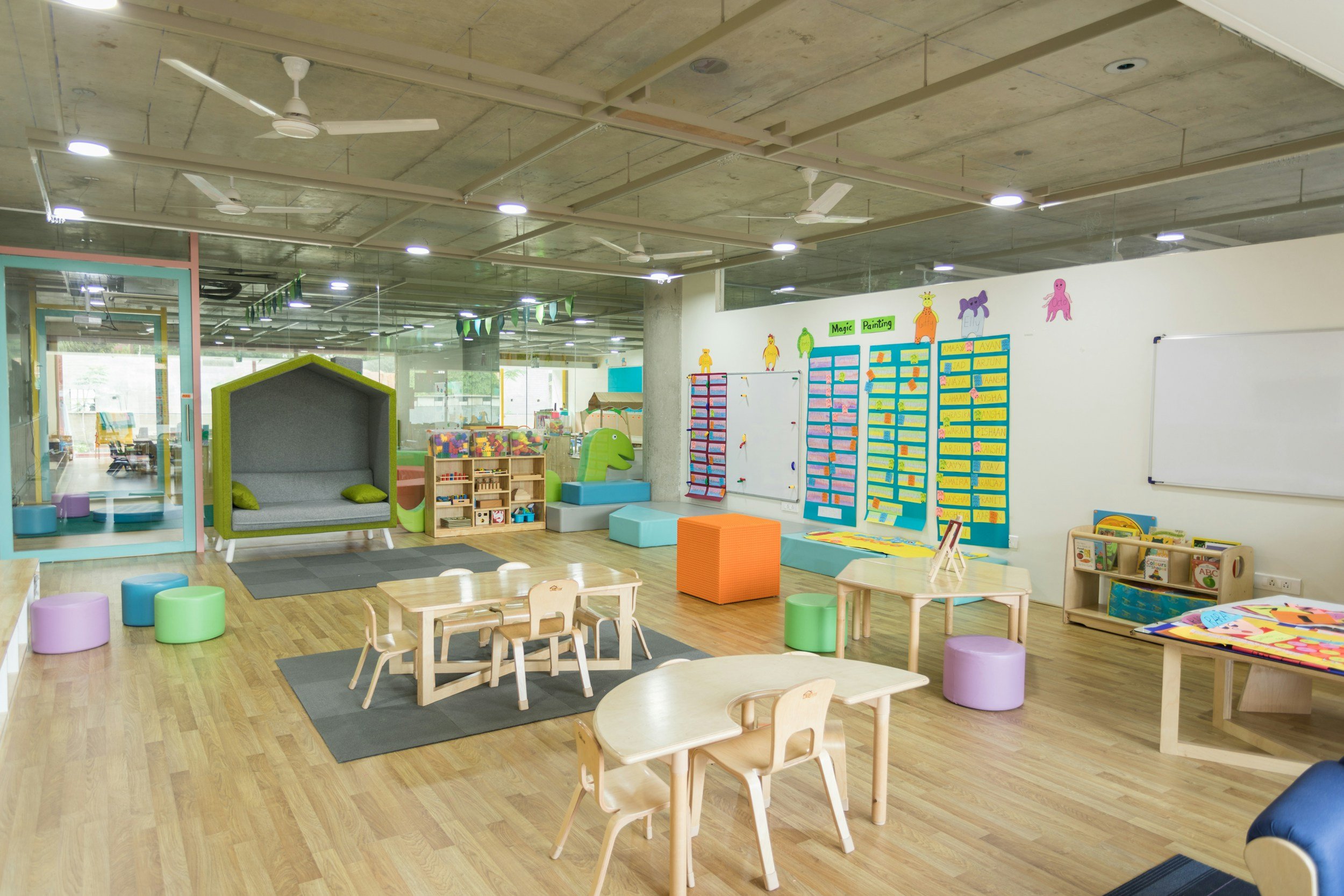 Bright and colorful children's playroom and learning area with small tables, chairs, cushioned seats, shelves of books and toys, and a whiteboard on the wall.