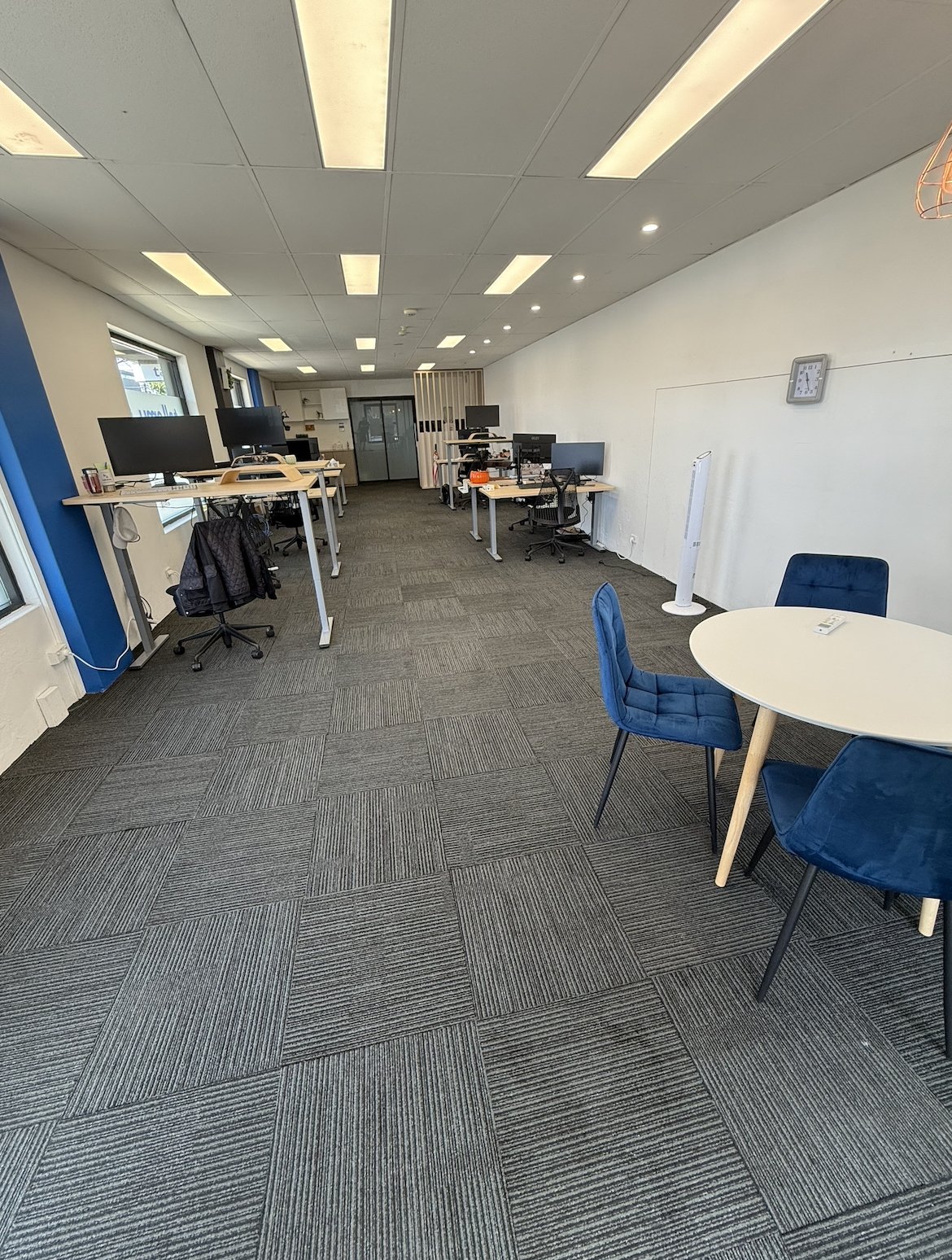 An open office space with multiple desks equipped with computer monitors, chairs, and various office supplies. There is a round table with three blue chairs, a standing fan, a wall clock, and large windows letting in natural light. The ceiling has rectangular light panels.