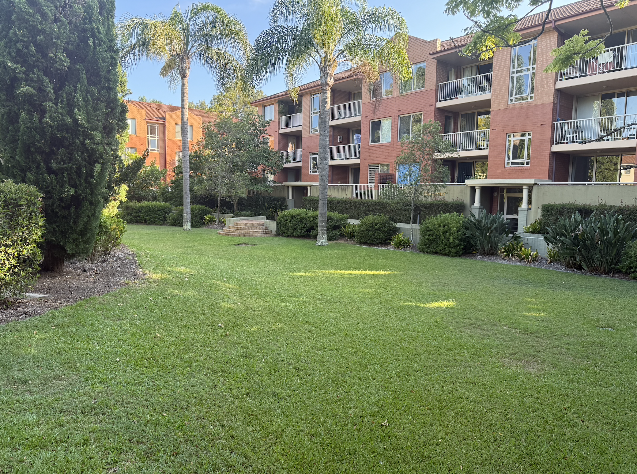 A well-maintained grassy courtyard with palm trees, bushes, and apartment building balconies in the background.