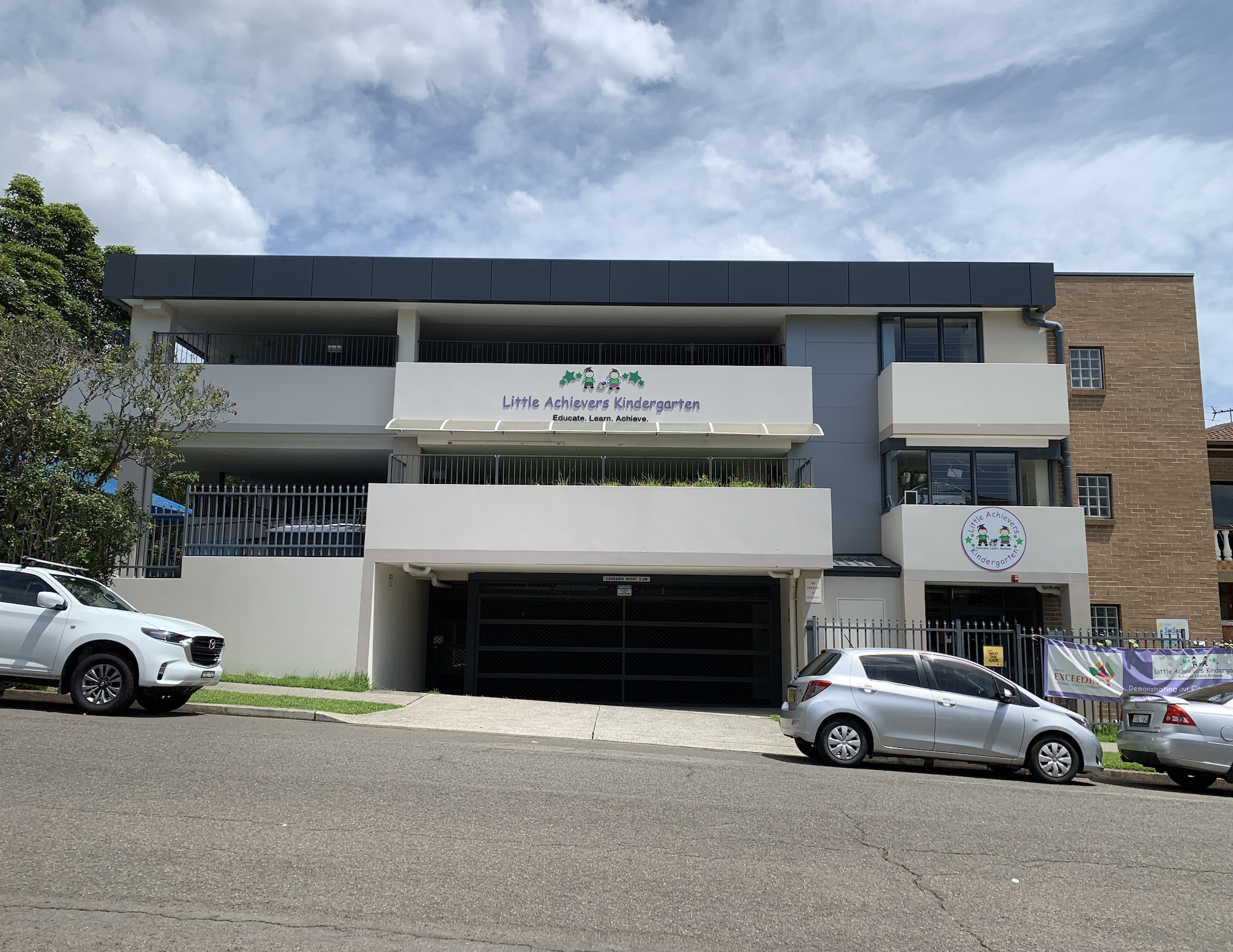 A three-story building with signs for Little Achievers Kindergarten, parked cars in front, and a cloudy sky above.