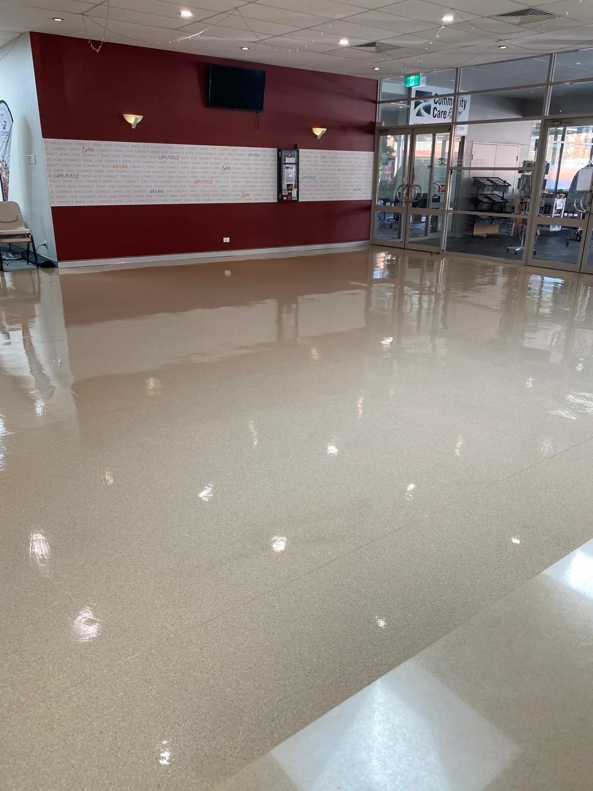 Empty indoor space with a glossy beige floor, red wall with white text, and glass doors leading to an outdoor area.