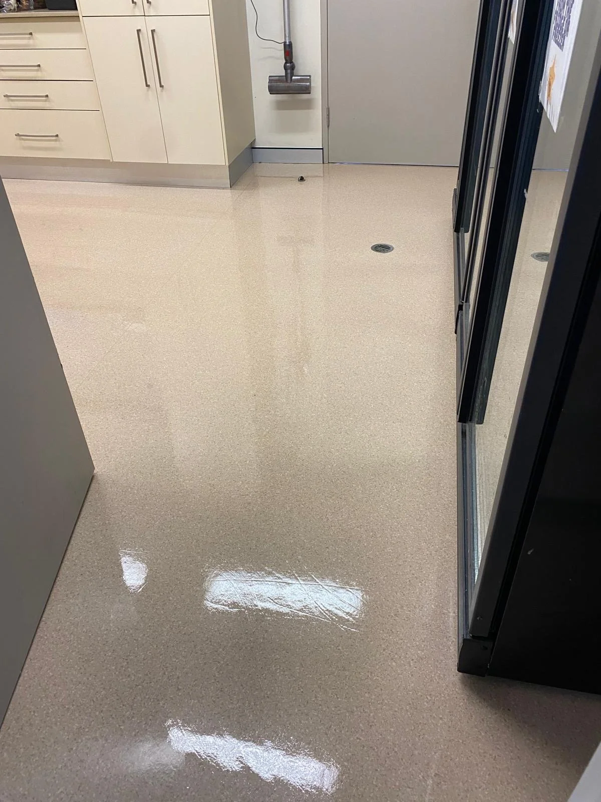 Clean, shiny beige tiled floor in a room with kitchen cabinets, a door in the background, and a refrigerator on the right