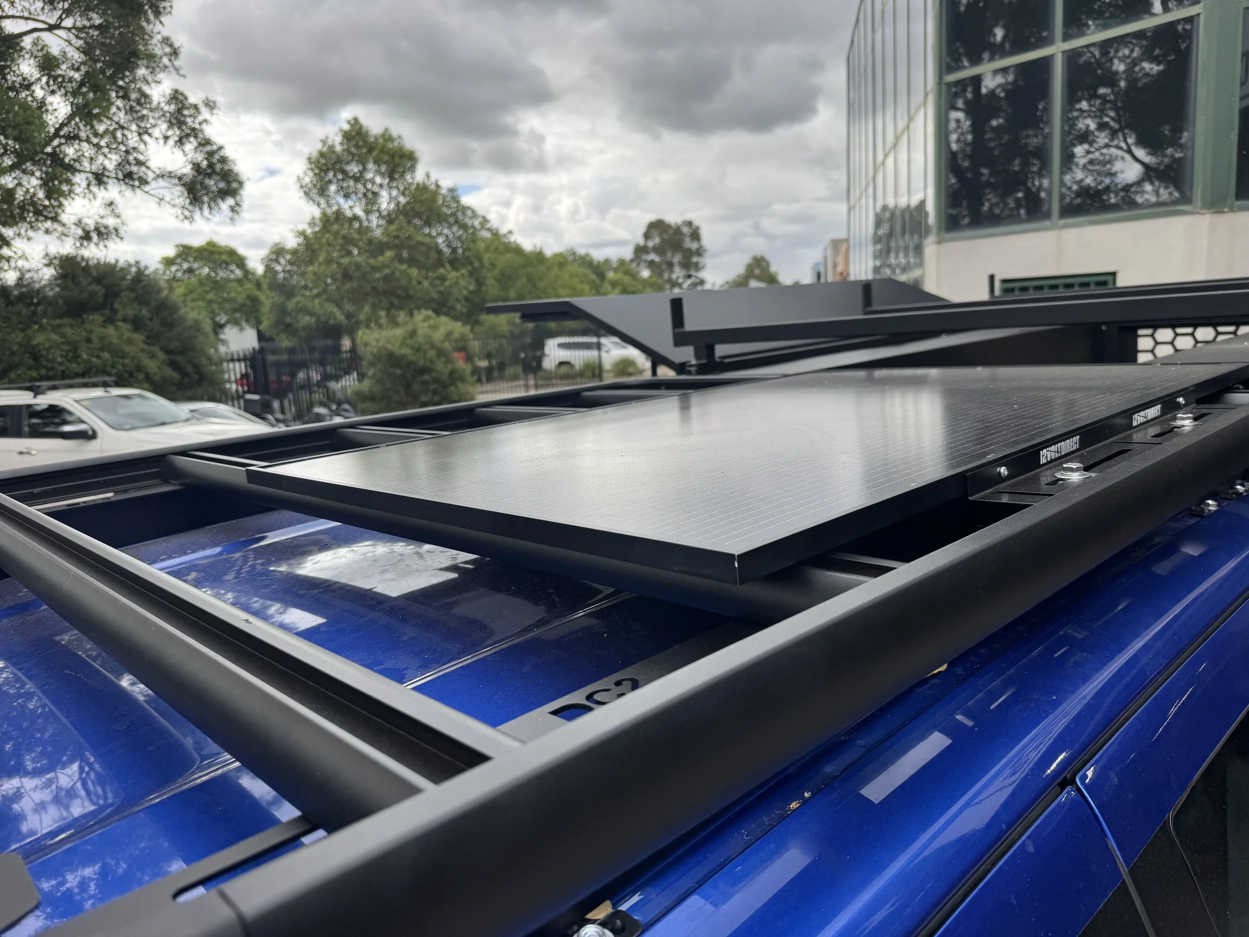 Close-up of a solar panel mounted on the roof of a blue vehicle with a modern building, trees, and a cloudy sky in the background.