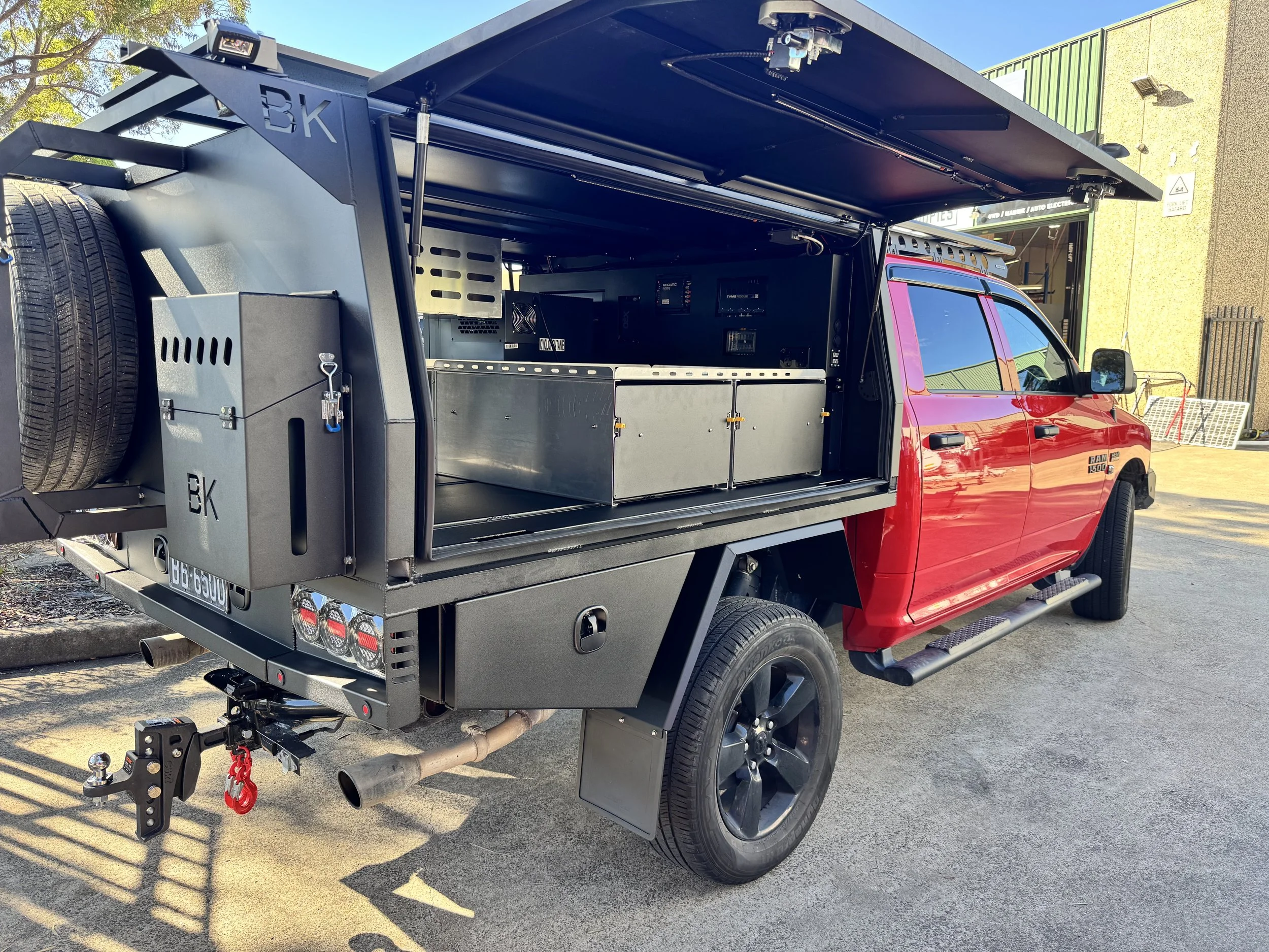 A red pickup truck with a black tool storage unit mounted in the truck bed, equipped with electrical and mechanical equipment, parked outside a building with a green and yellow exterior and a sign that says 'US'.