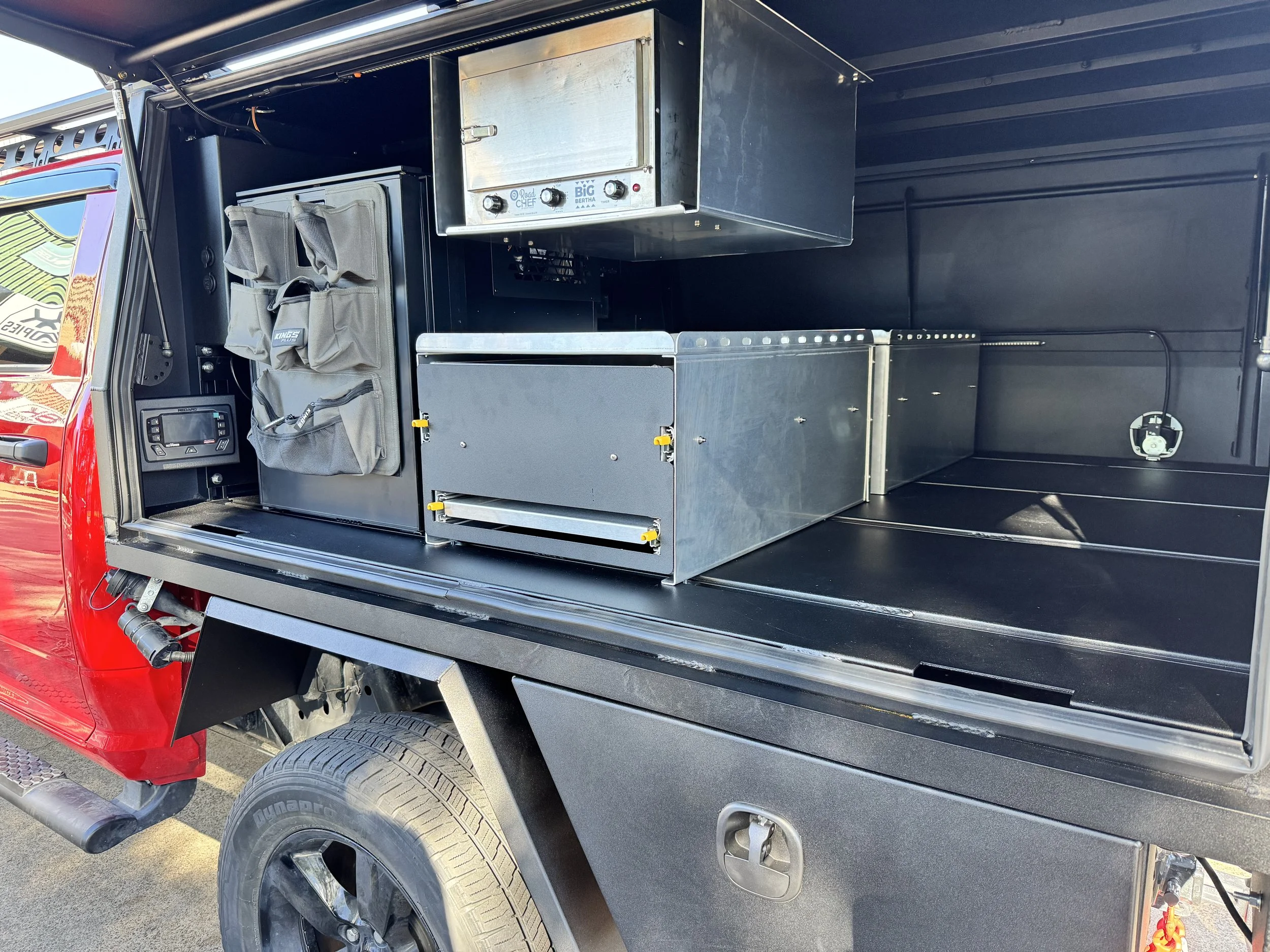 The image shows the open side of a utility truck with various compartments. There are tools and equipment, including a control panel labeled 'Road Chef' and 'Big Bertha,' and organized storage pouches. The truck has a black interior with metallic and
