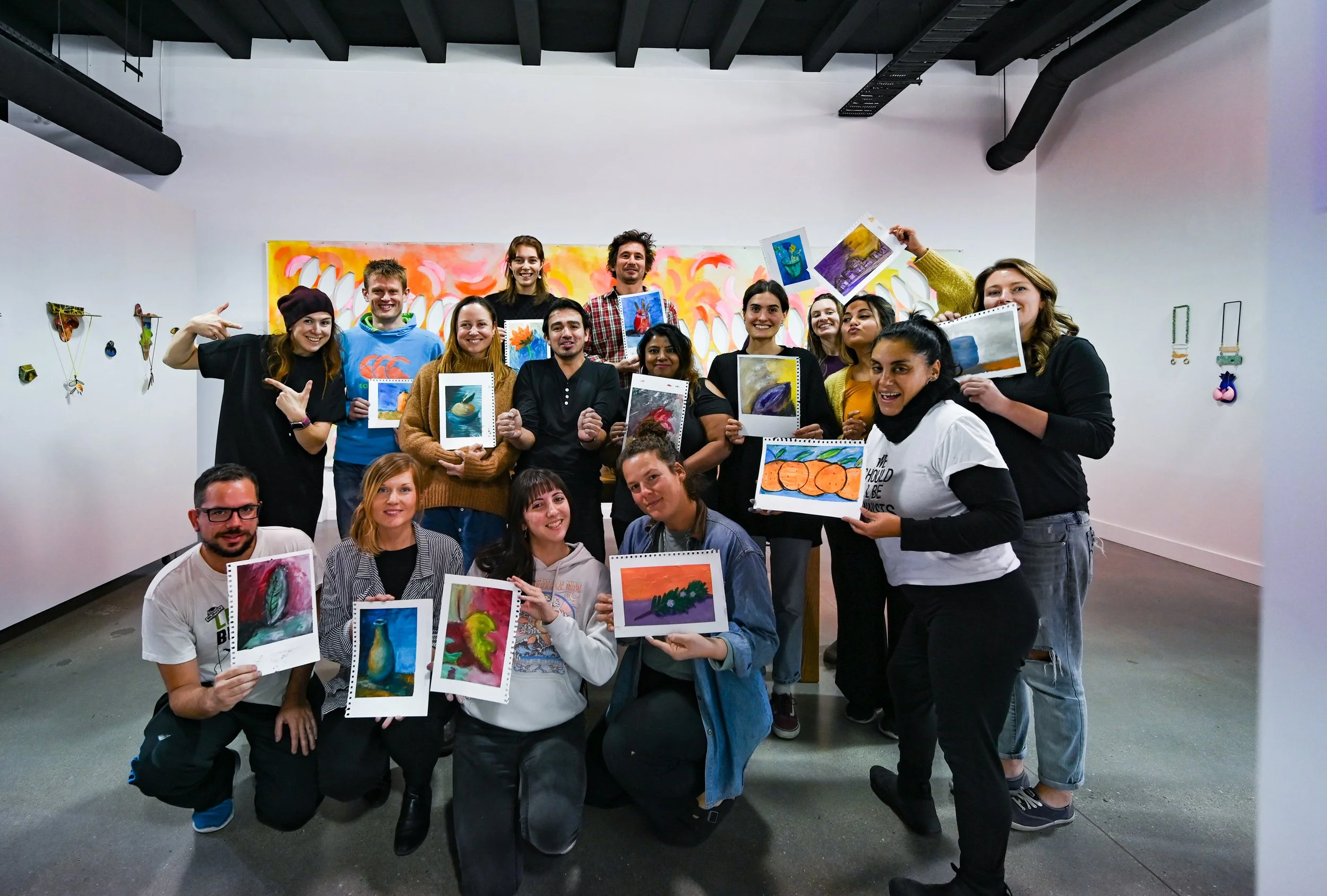 A diverse group of people in an art gallery holding colorful paintings and smiling, with abstract artwork on the wall behind them.
