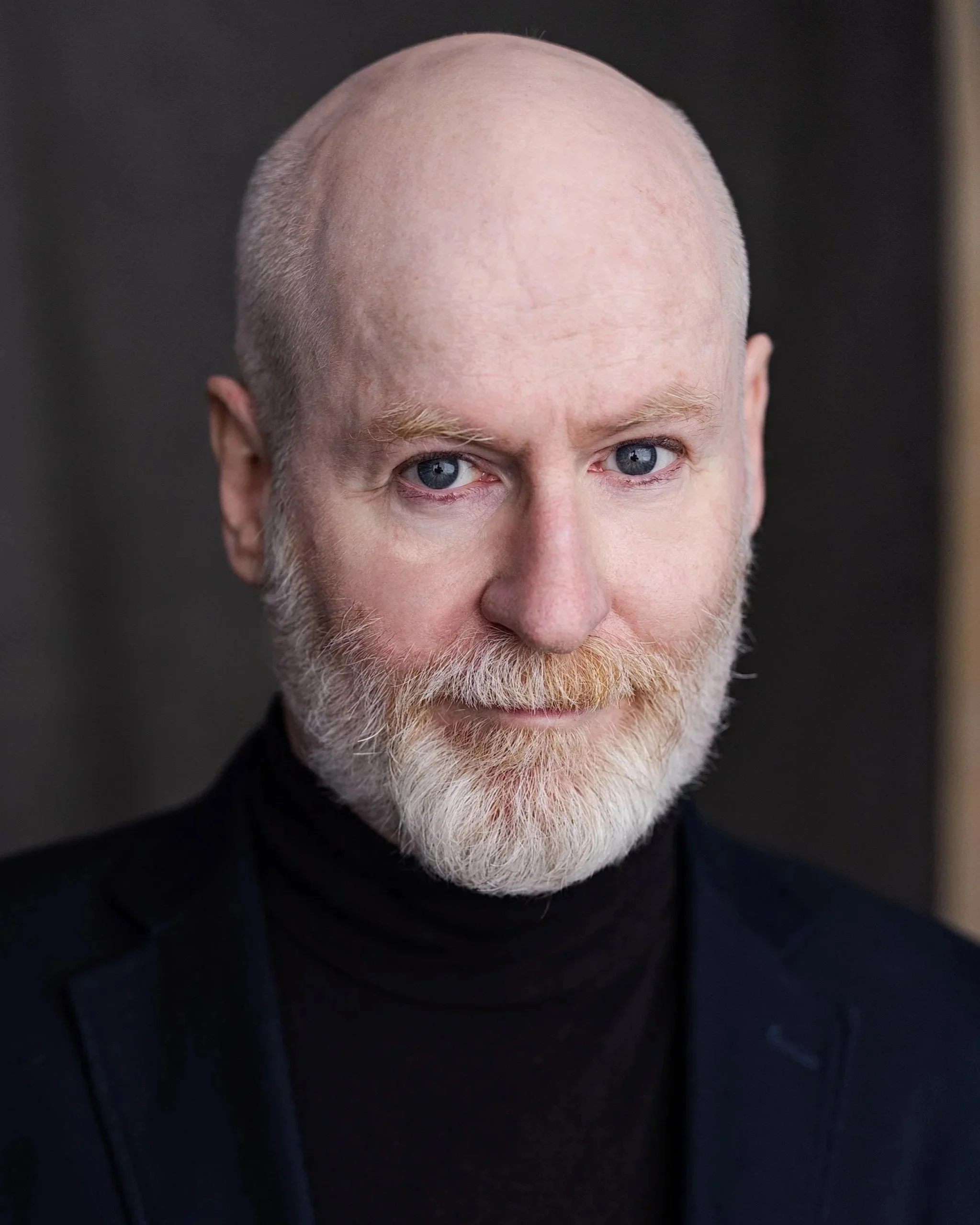 A bearded man in a turtleneck and blazer with a blurred gray background.