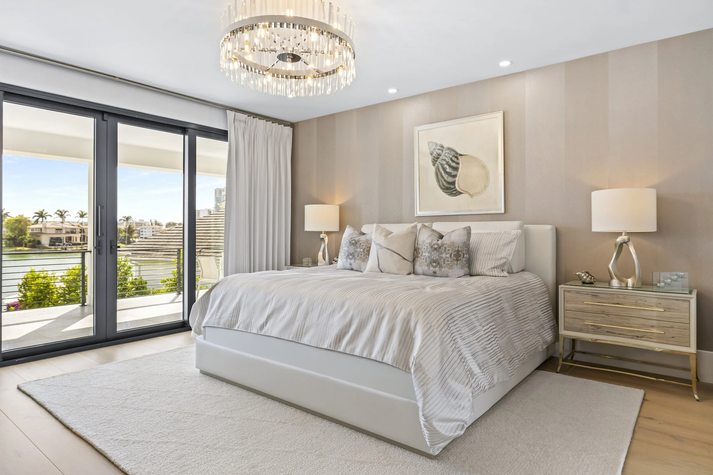 Bright bedroom with a large white bed, decorative pillows, cream-colored walls, artwork above the bed, two nightstands with lamps, sliding glass door leading to a balcony, and a chandelier on the ceiling.