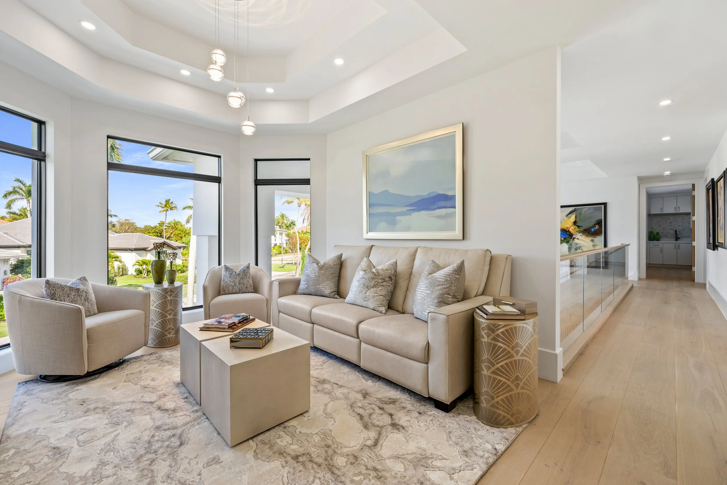 Bright living room with a beige sofa, two armchairs, a coffee table, large windows with a view of a sunny yard and palm trees, contemporary art on the walls, light wood flooring, and modern ceiling lighting.