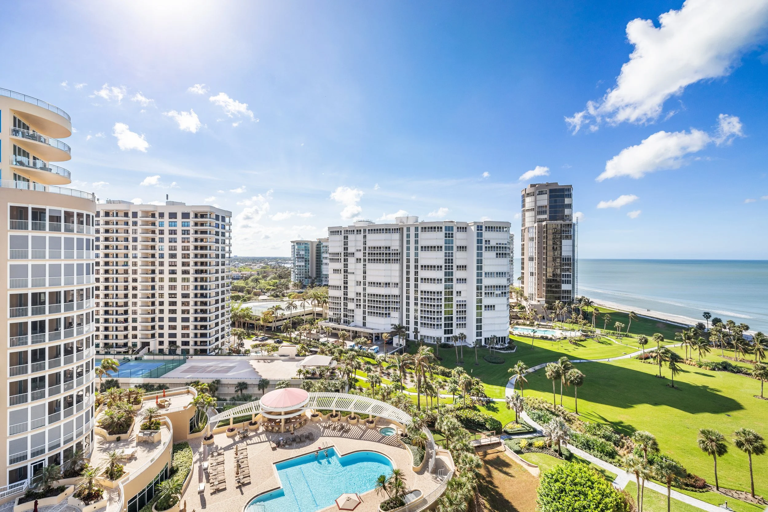 High-rise buildings near a beach with a pool and green lawn, under a bright blue sky with some clouds.
