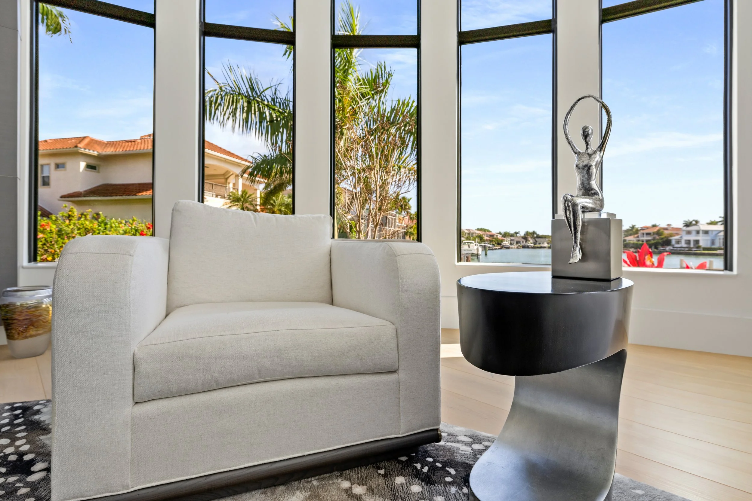 Modern living room with white sofa, black side table with a silver sculpture of a woman sitting gracefully with arms raised, large windows showing palm trees, a canal, and neighboring houses.