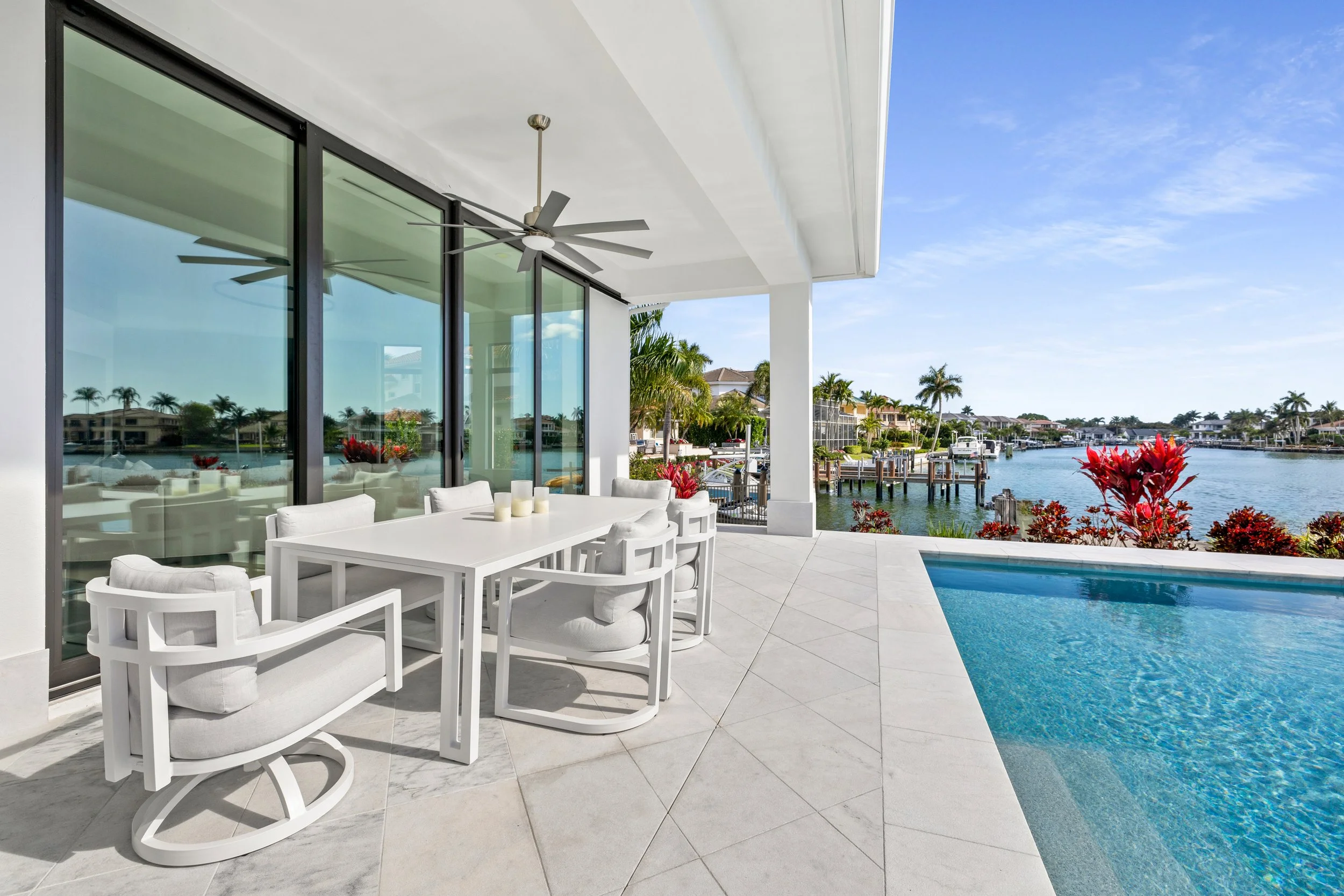 A balcony with white furniture and a swimming pool overlooking a canal with boats and houses, surrounded by palm trees and colorful plants.