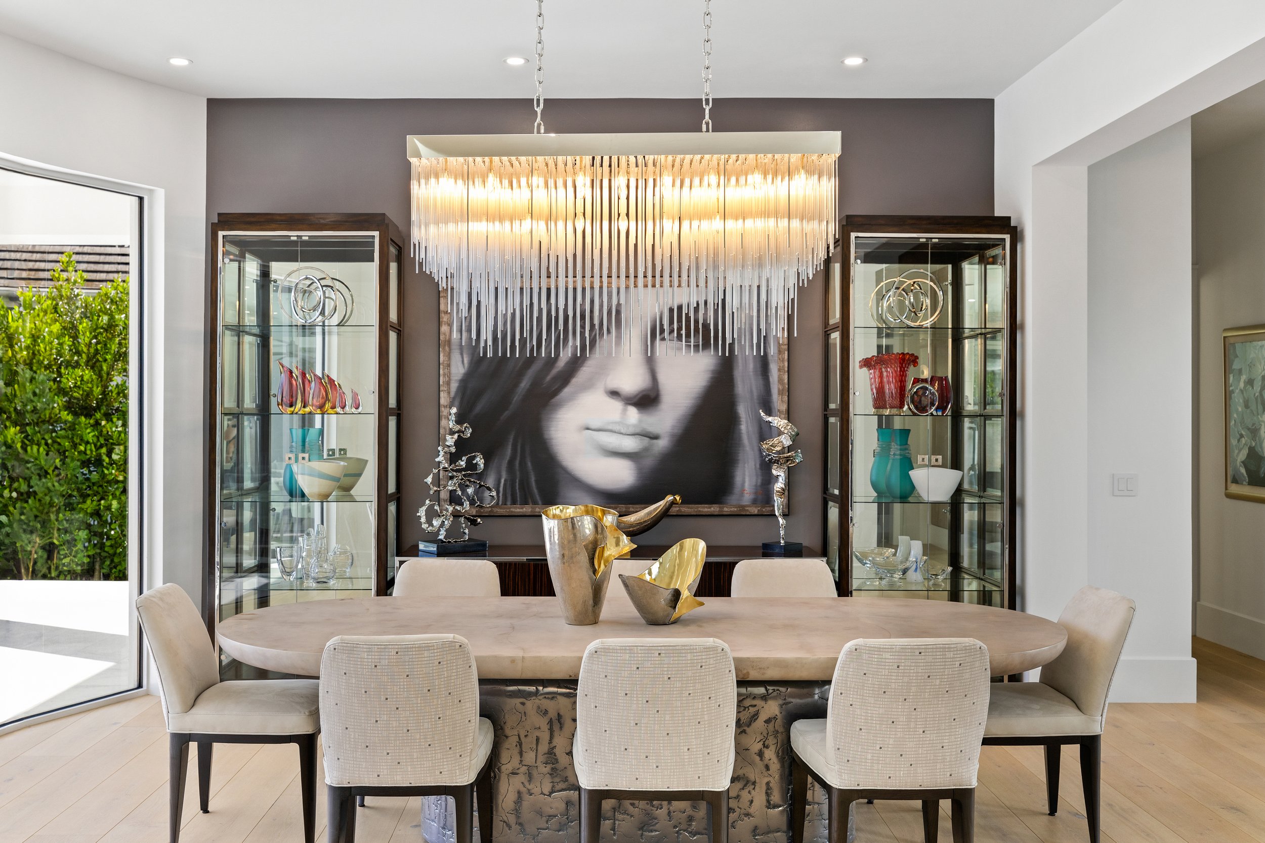 A dining room with a large oval table, eight beige upholstered chairs, a black-and-white portrait of a woman's face on the wall, a modern chandelier hanging above, and glass display cabinets filled with decorative items. There's a sliding glass door to the outside showing greenery.
