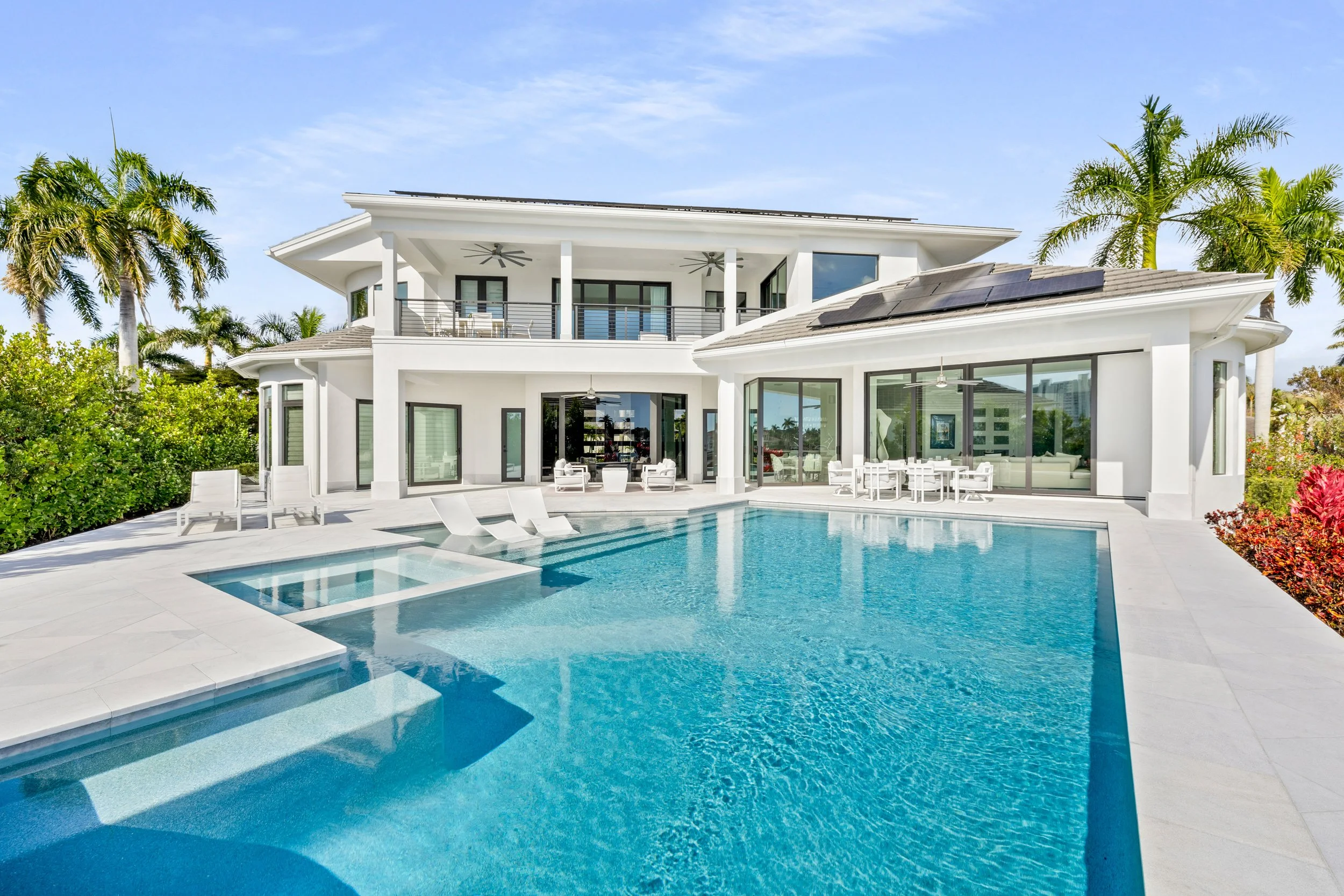 Modern white house with large swimming pool, surrounded by tropical trees and plants, under a clear blue sky.