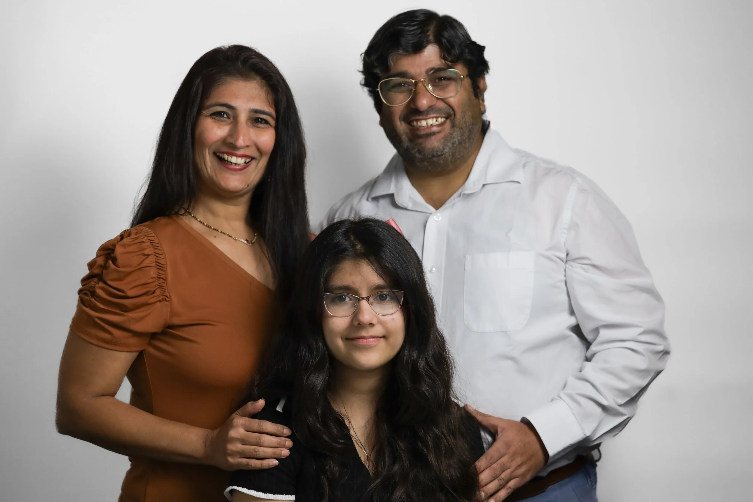 A family of three, a woman, a man, and a girl, posing together indoors against a plain background. The woman has long dark hair and is wearing a brown top. The girl has long curly dark hair, glasses, and is wearing a black top. The man has curly dark