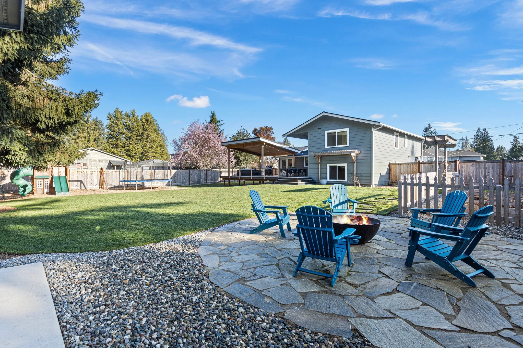 Backyard with green lawn, stone patio, fire pit surrounded by blue Adirondack chairs, wooden fence, house, trees, and children's play area with slides and a trampoline, under a clear blue sky.