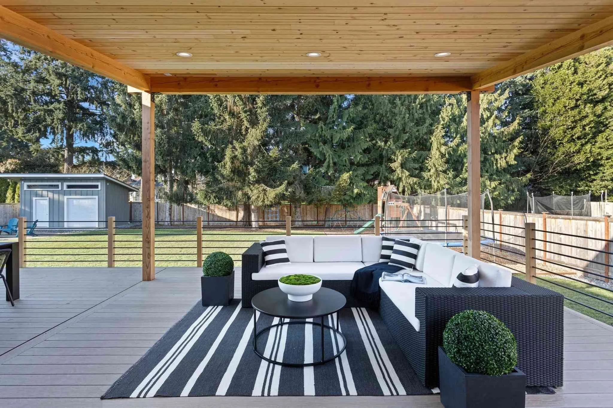 Outdoor covered patio with black wicker sectional sofa with white cushions, black and white striped pillows, black round coffee table with a white bowl, two square black planters with green shrubs, striped outdoor rug, and a fenced backyard with trees, grass, a shed, and a playset.