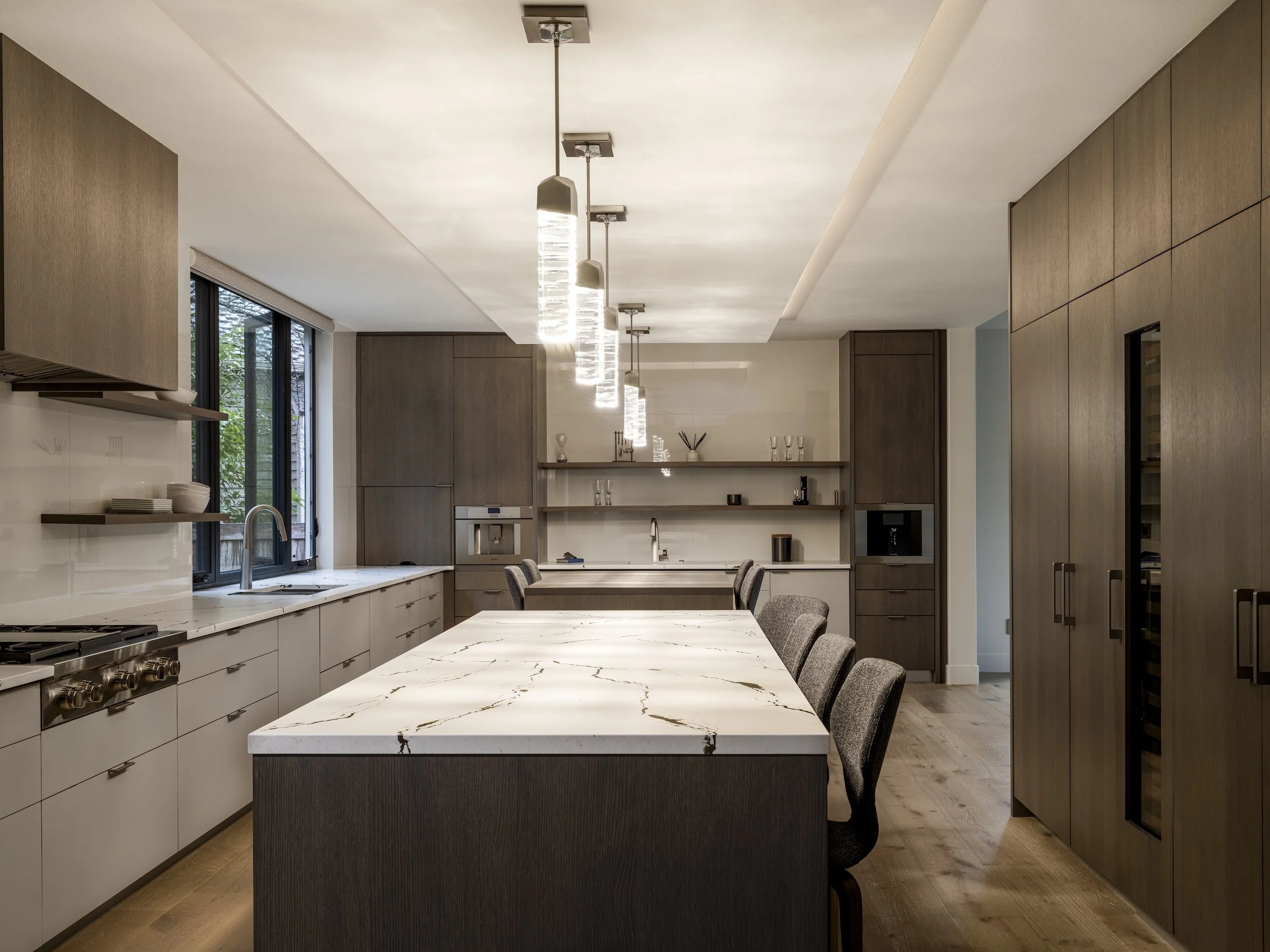 Modern kitchen with large marble island, gray chairs, wood cabinets, and pendant lighting.
