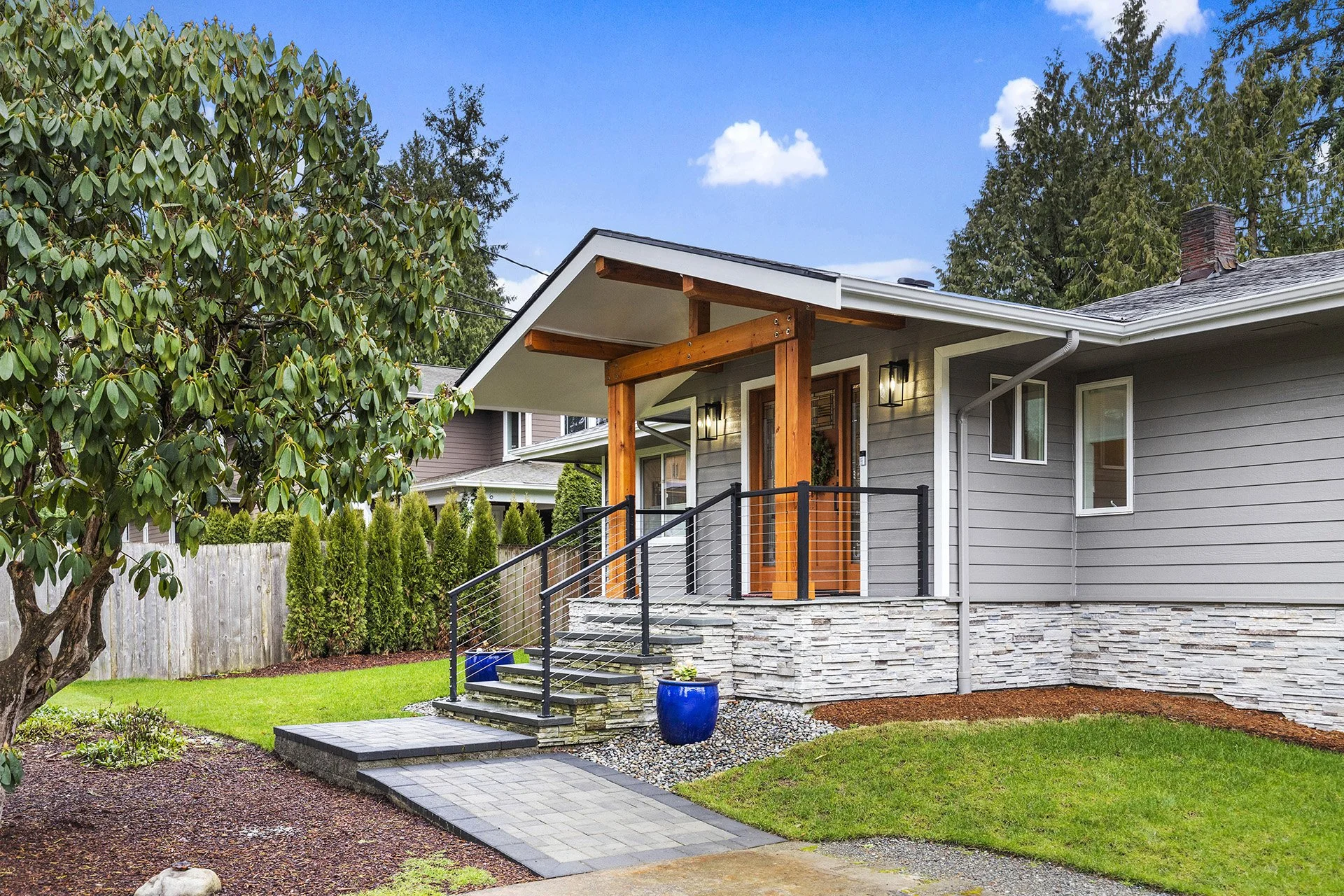 Front of a modern house with a small porch, wooden columns, a black metal railing, and stairs leading to the door. The house has gray siding, a stone foundation, and outdoor lighting. There are trees and a landscaped yard with grass, a paved walkway, and a blue pot with plants.