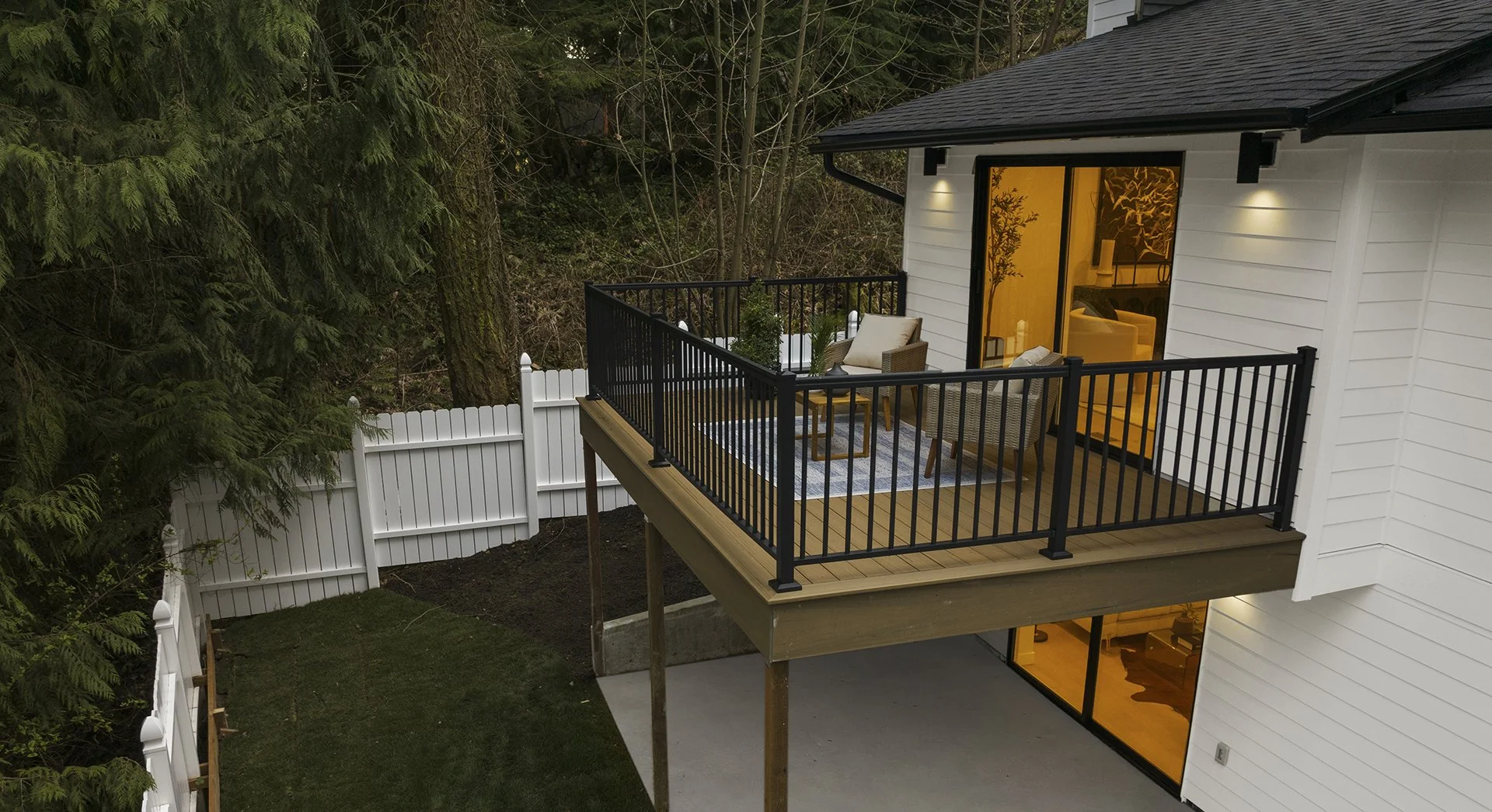 A balcony with black railing attached to a white house, overlooking a backyard with grass and tall trees, with interior lighting visible through large windows and sliding glass door.