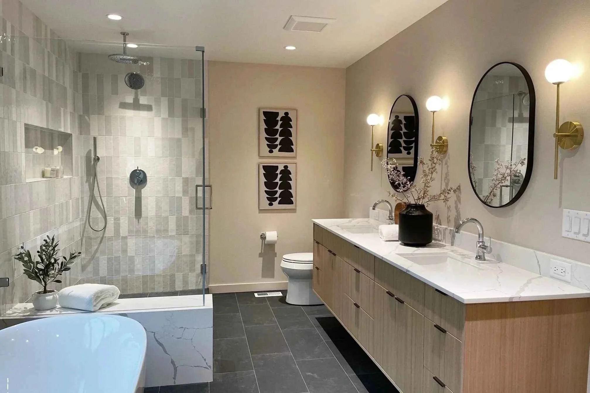 Modern bathroom with dual sinks, large oval mirrors, wall-mounted sconces, a shower with glass enclosure, a toilet, and minimalistic decor including a vase with branches and framed abstract art.