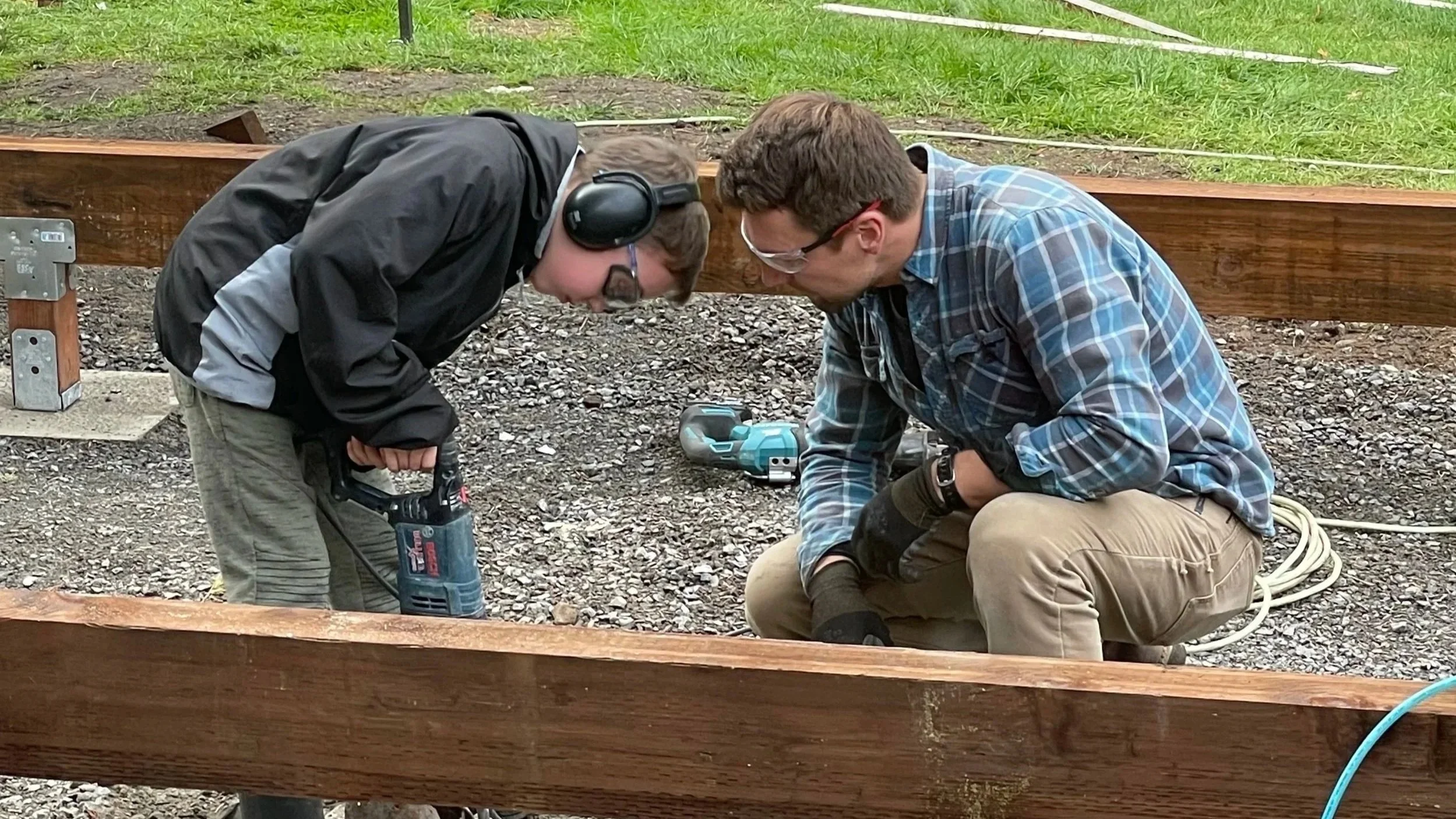 Two men working outdoors with power tools, one holding a drill and the other watching, surrounded by wood and construction equipment.