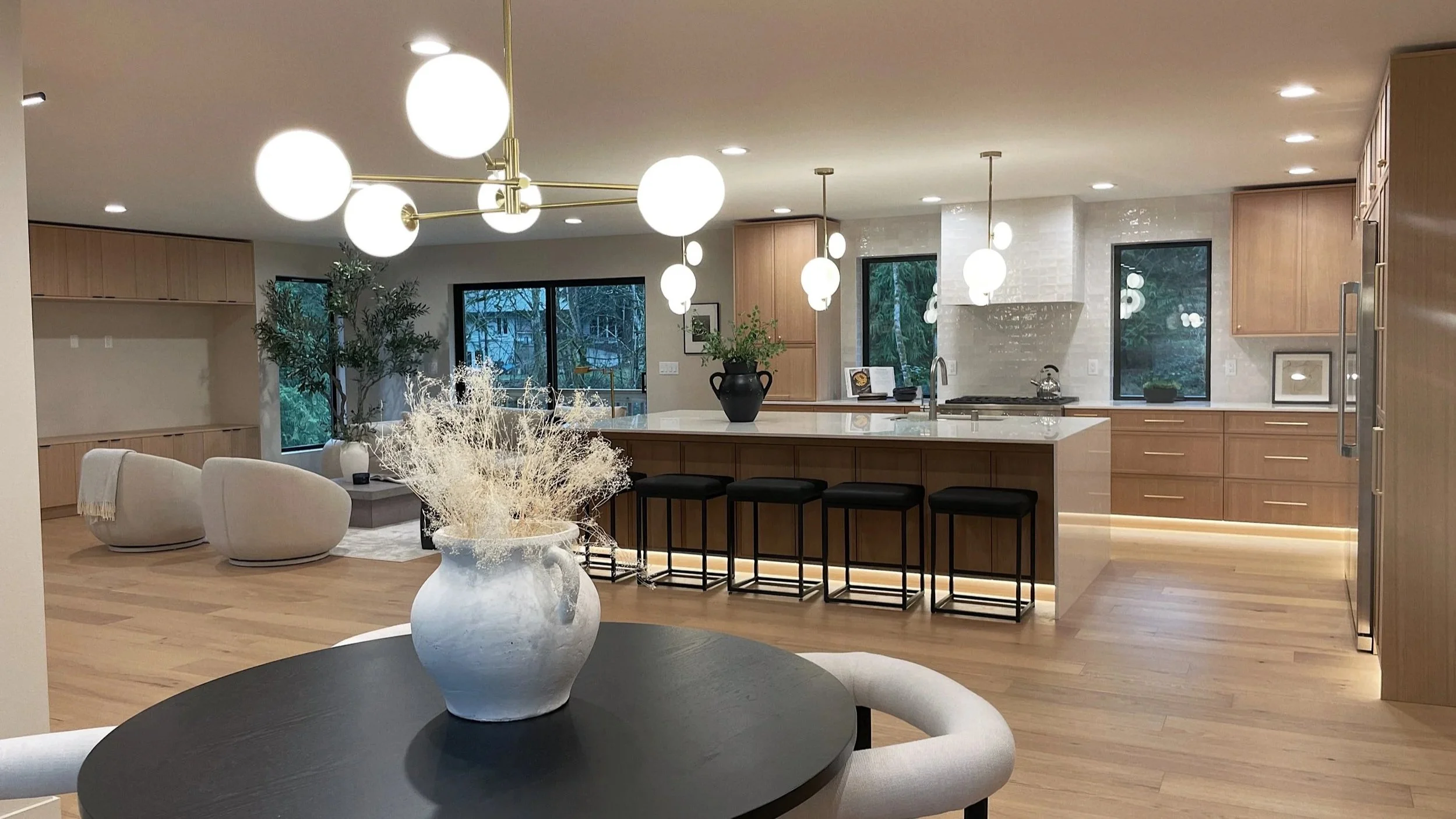 Modern open-concept kitchen and living room with wooden cabinets, large island, bar stools, and contemporary lighting fixtures. Decor includes a potted plant, vases with dried flowers, and large windows with greenery outside.